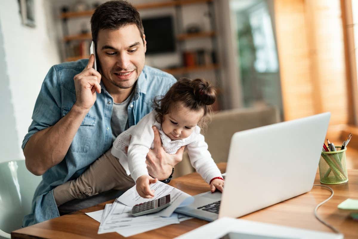 Man on a laptop talking on a phone with a baby reaching for a calculator in his other arm
