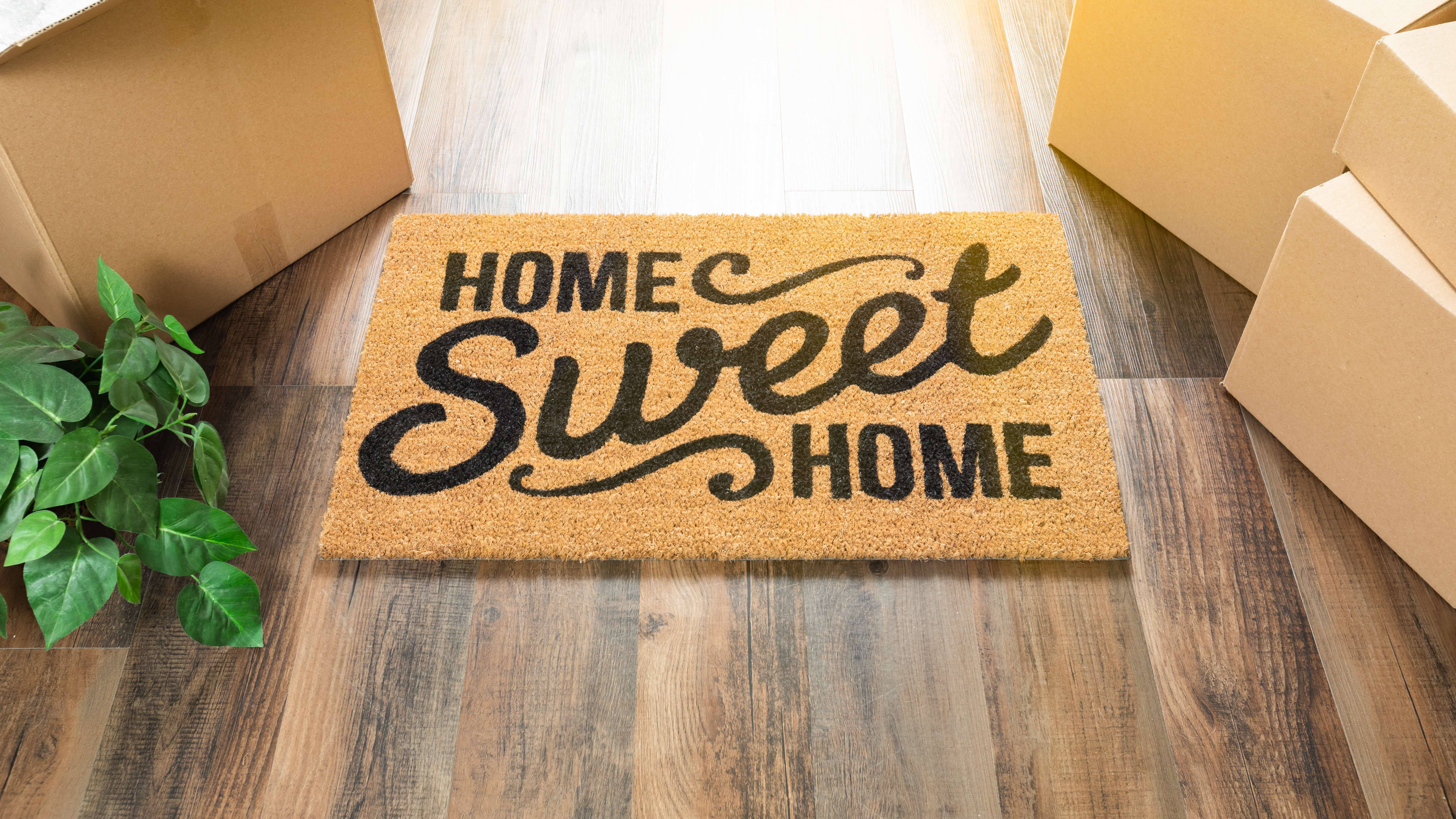 A hessian door mat which has the words 'Home sweet home' printed on it sits on the floor of a sunny hallway, surrounded by cardboard boxes and a house plant