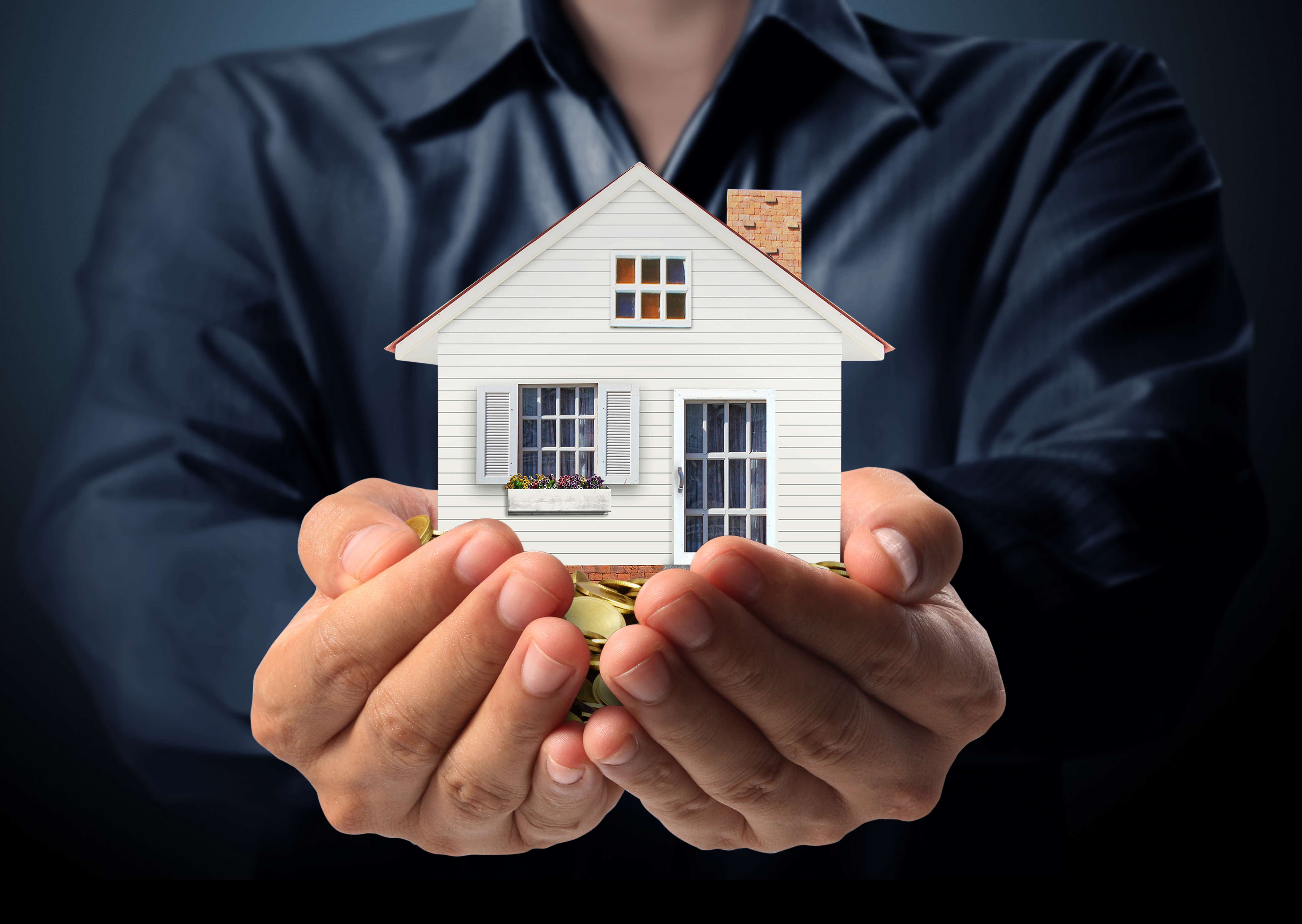 Close up of a male torso wearing a dark blue shirt, arms outstretched holding a model of a house, which is sitting on top of a pile of coins