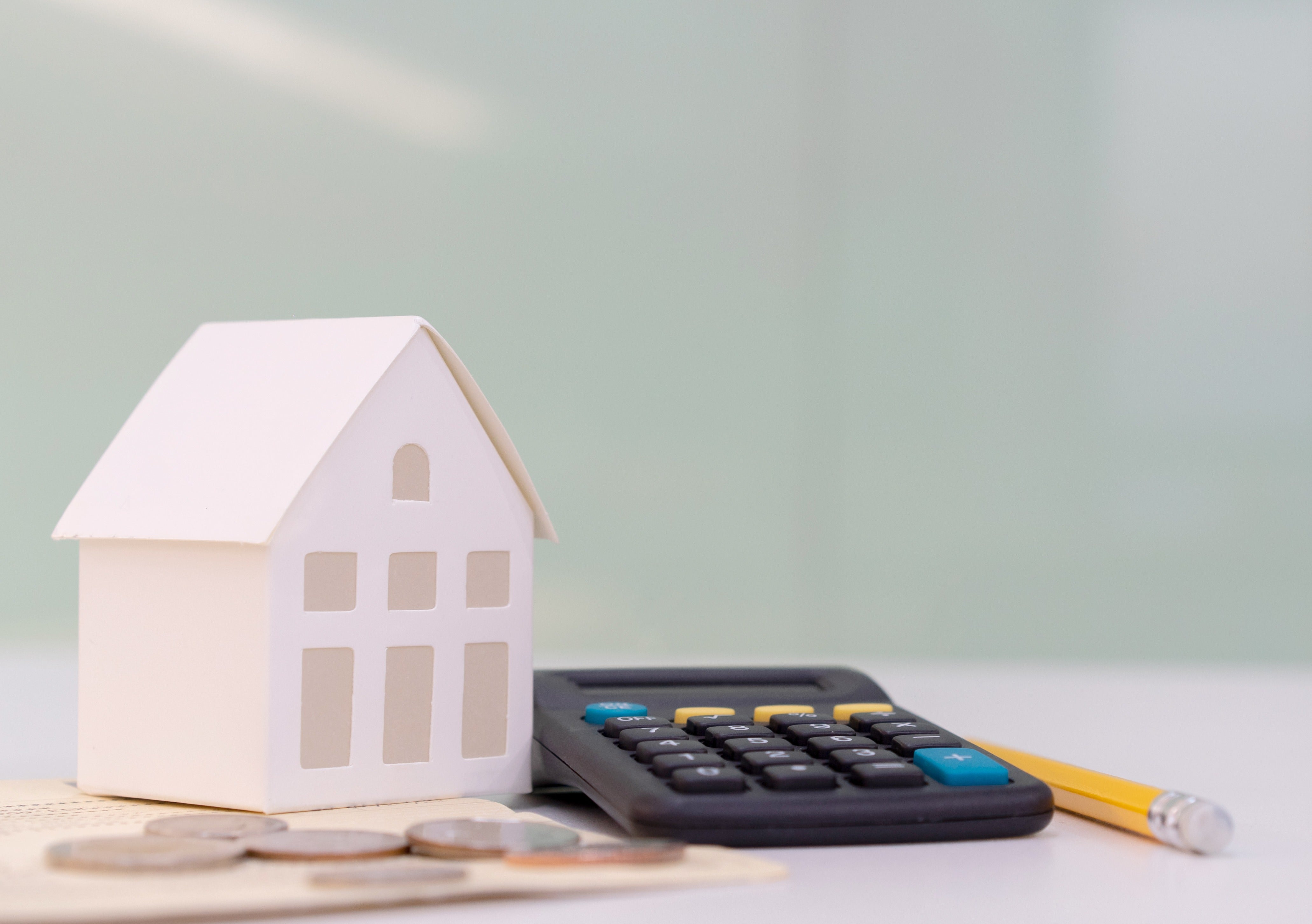 On a desk is a small white model of a house, a calculator, a pencil and some coins