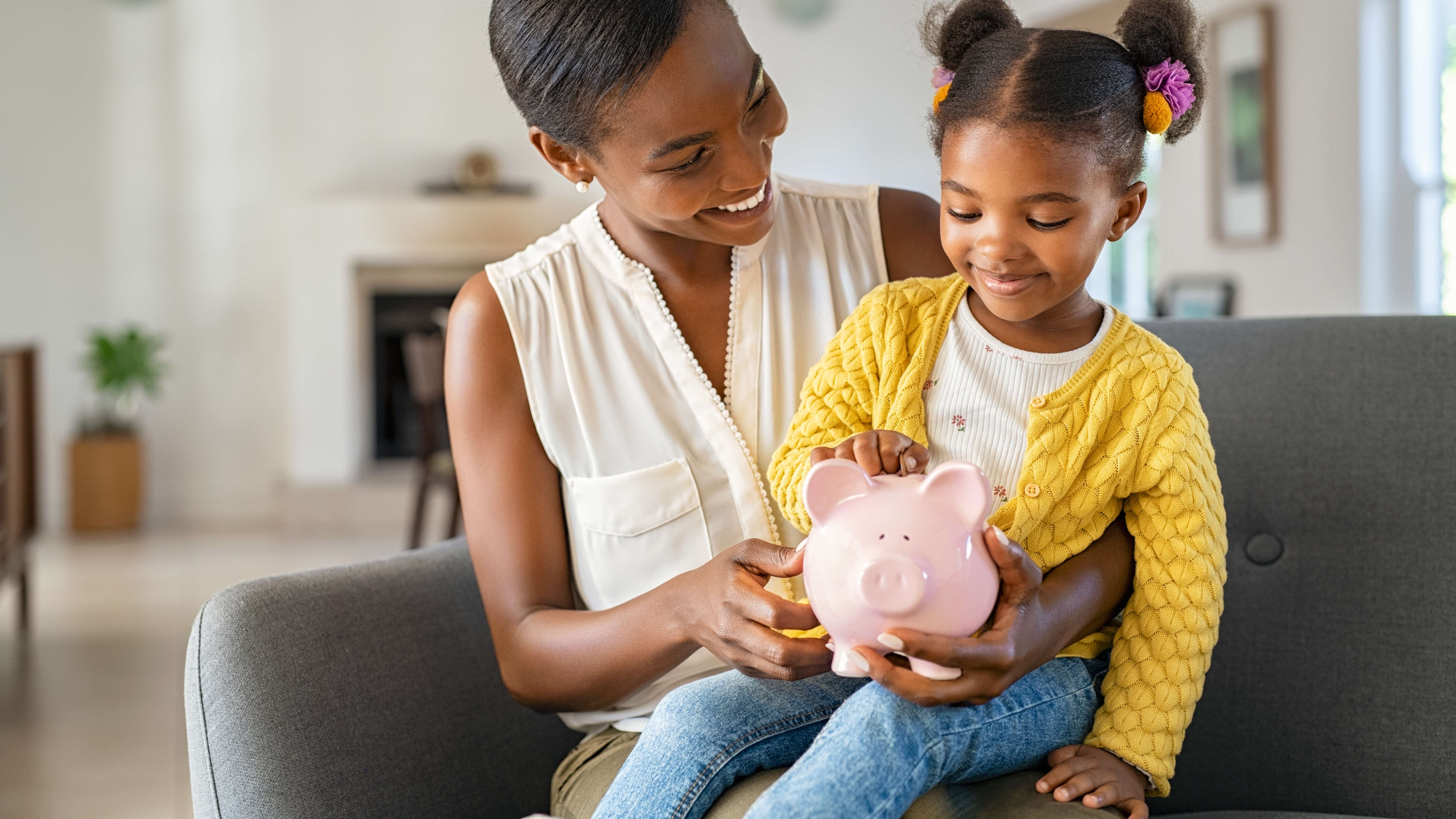 A black lady sits on a sofa in a large living room with a young girl on her lap. The girl is around 6 years old with bunches in her hair and is holding a pink pig money bank. She is putting a coin into the bank and both are smiling