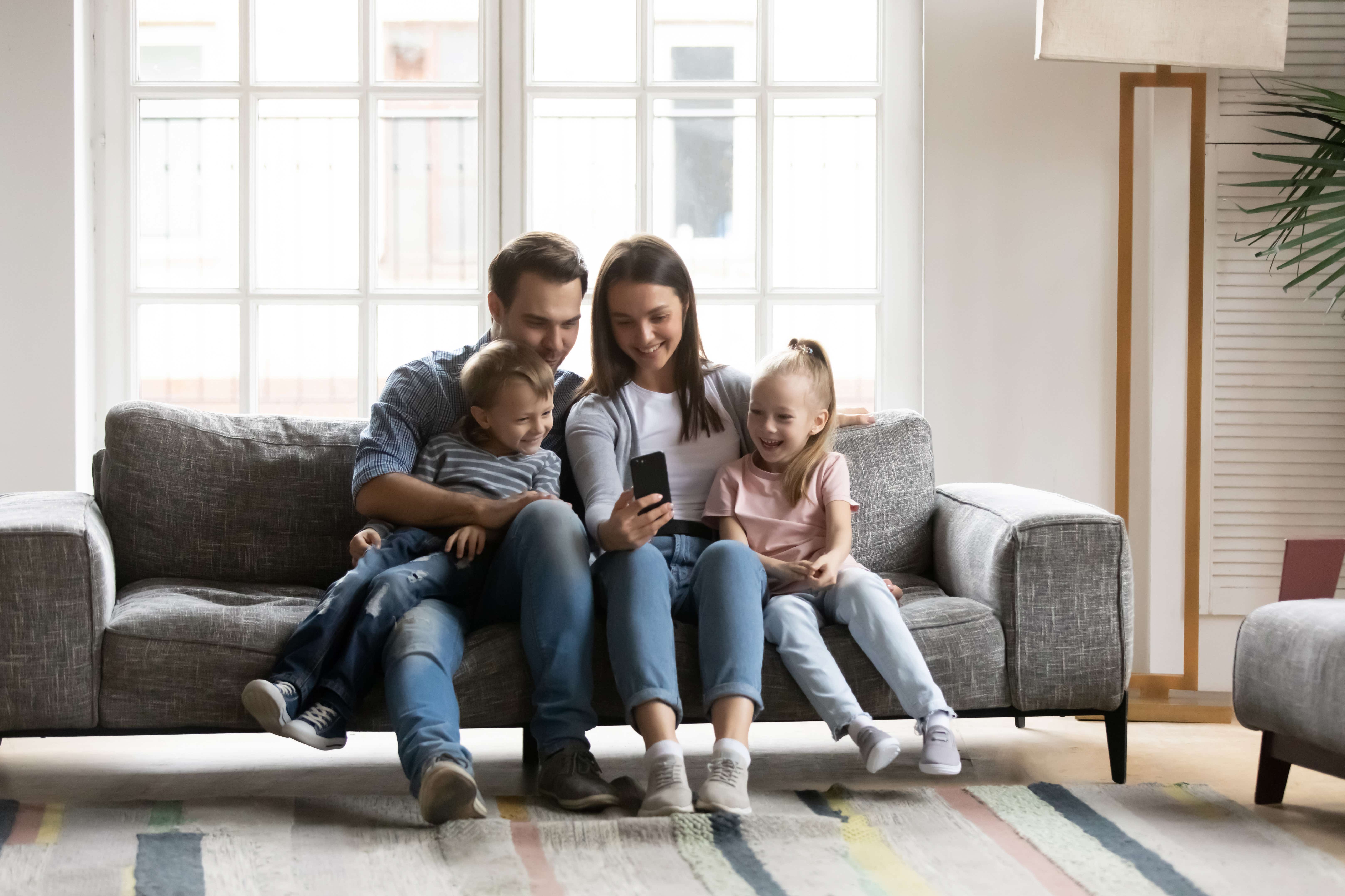 A young family with two children sit on a small grey sofa in the centre of their living room, there is a large window behind them and a rug at their feet