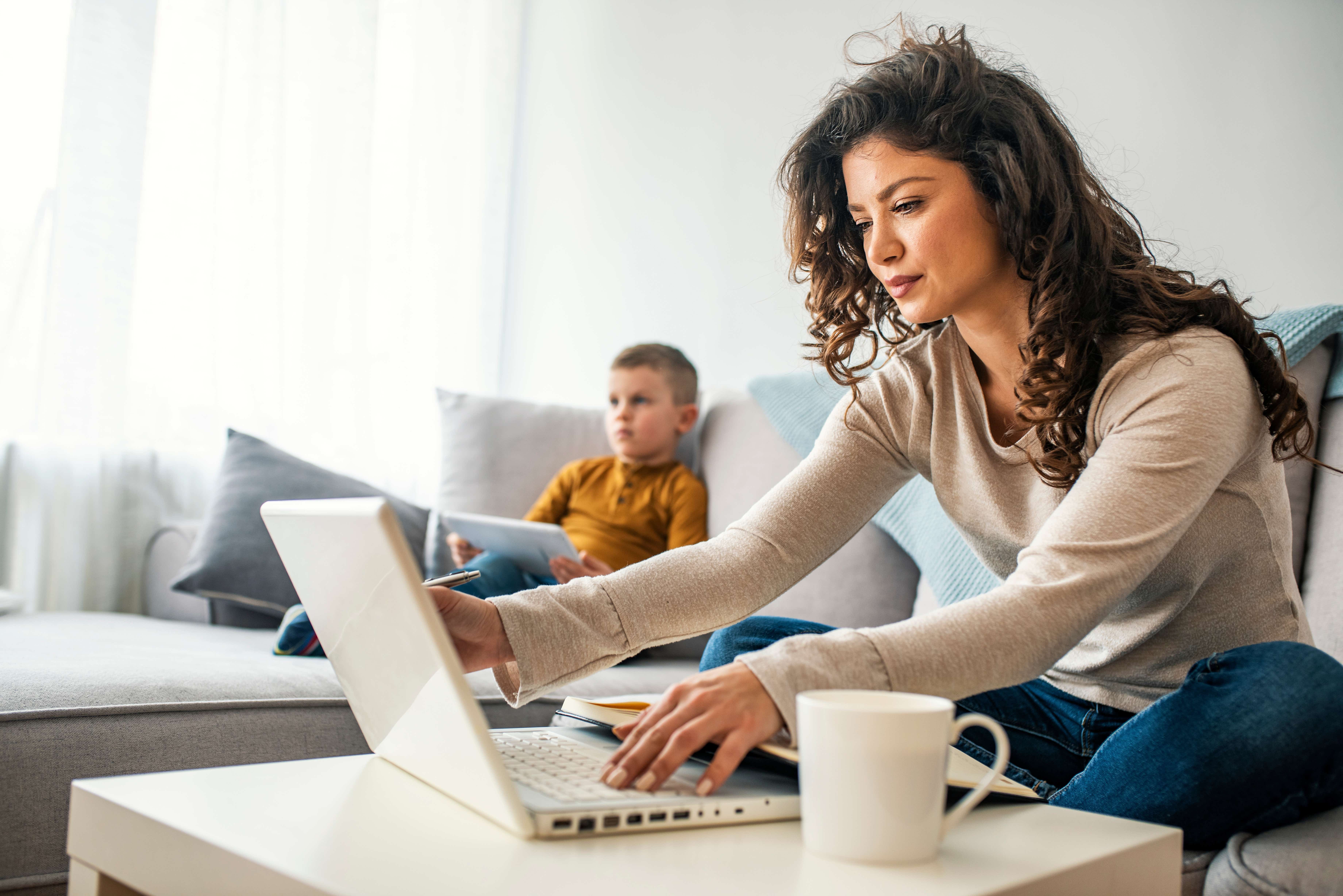 A young woman sits on the sofa with a laptop open on the coffee table in front of her. She is holding and looking at the screen with a slight smile. In the background, her young son, also sitting on the sofa, is using a tablet.