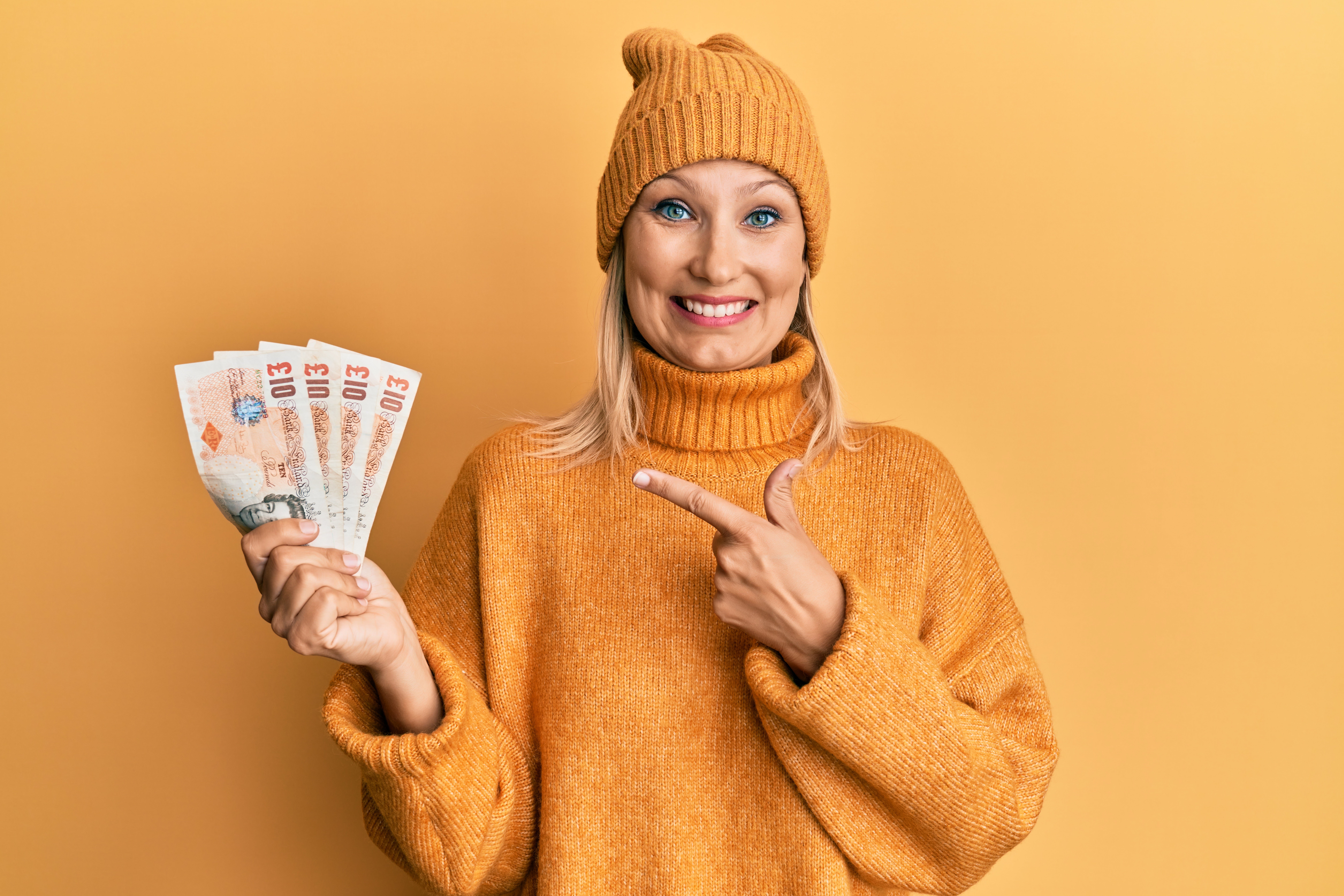 A young white woman dressed entirely in mustard yellow and standing against a mustard yellow backdrop holds a fan of UK bank notes in her right hand. He left hand is pointing to the money and she has a big smile on her face. 