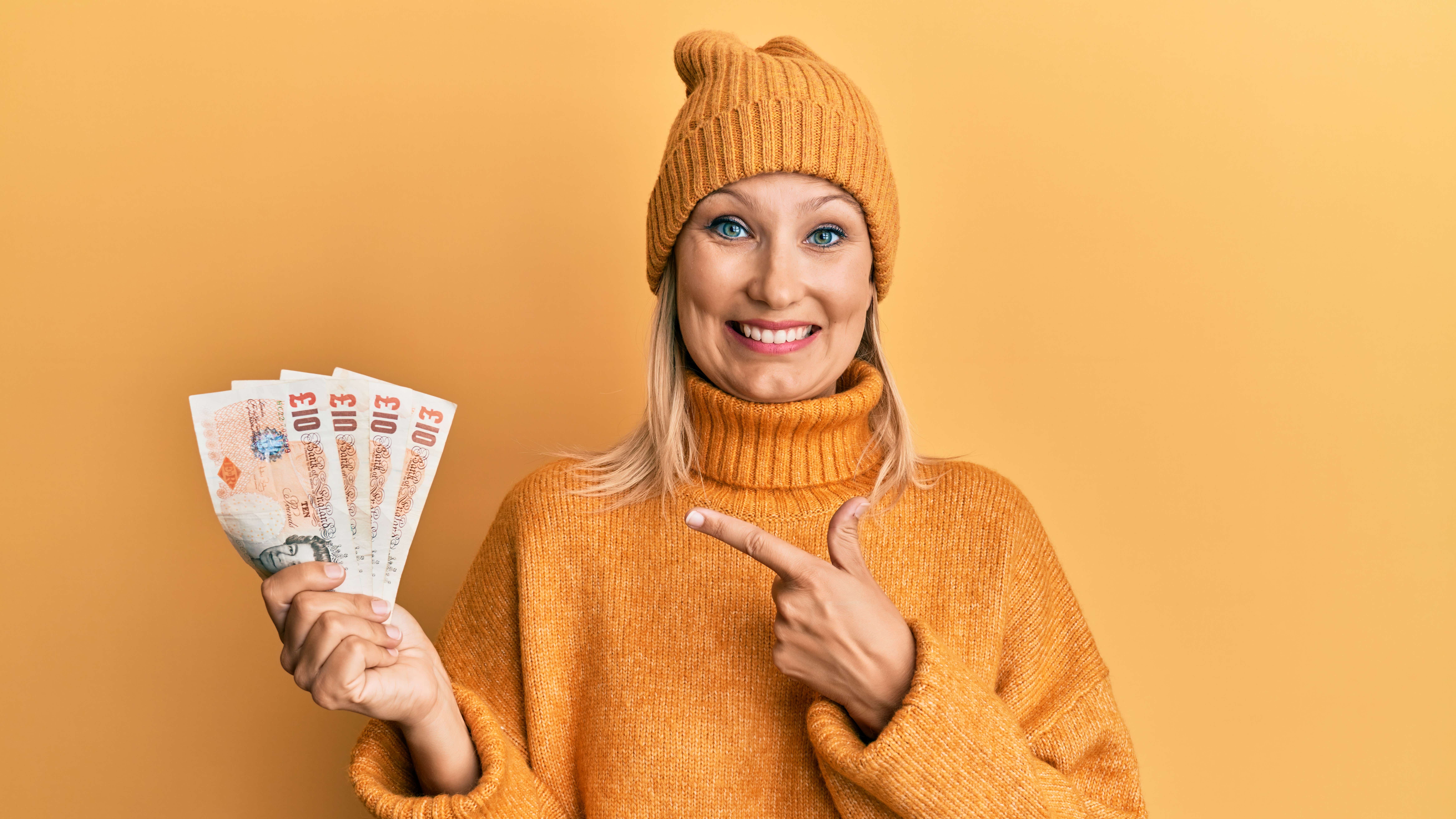 A young white woman dressed entirely in mustard yellow and standing against a mustard yellow backdrop holds a fan of UK bank notes in her right hand. He left hand is pointing to the money and she has a big smile on her face.
