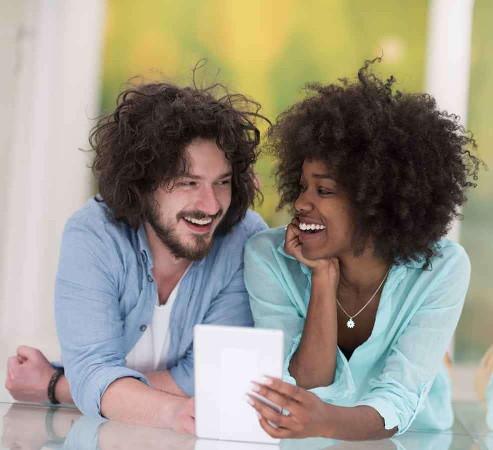 A young multi-ethnic couple look at each other with large smiles, they are jointly holding a tablet device