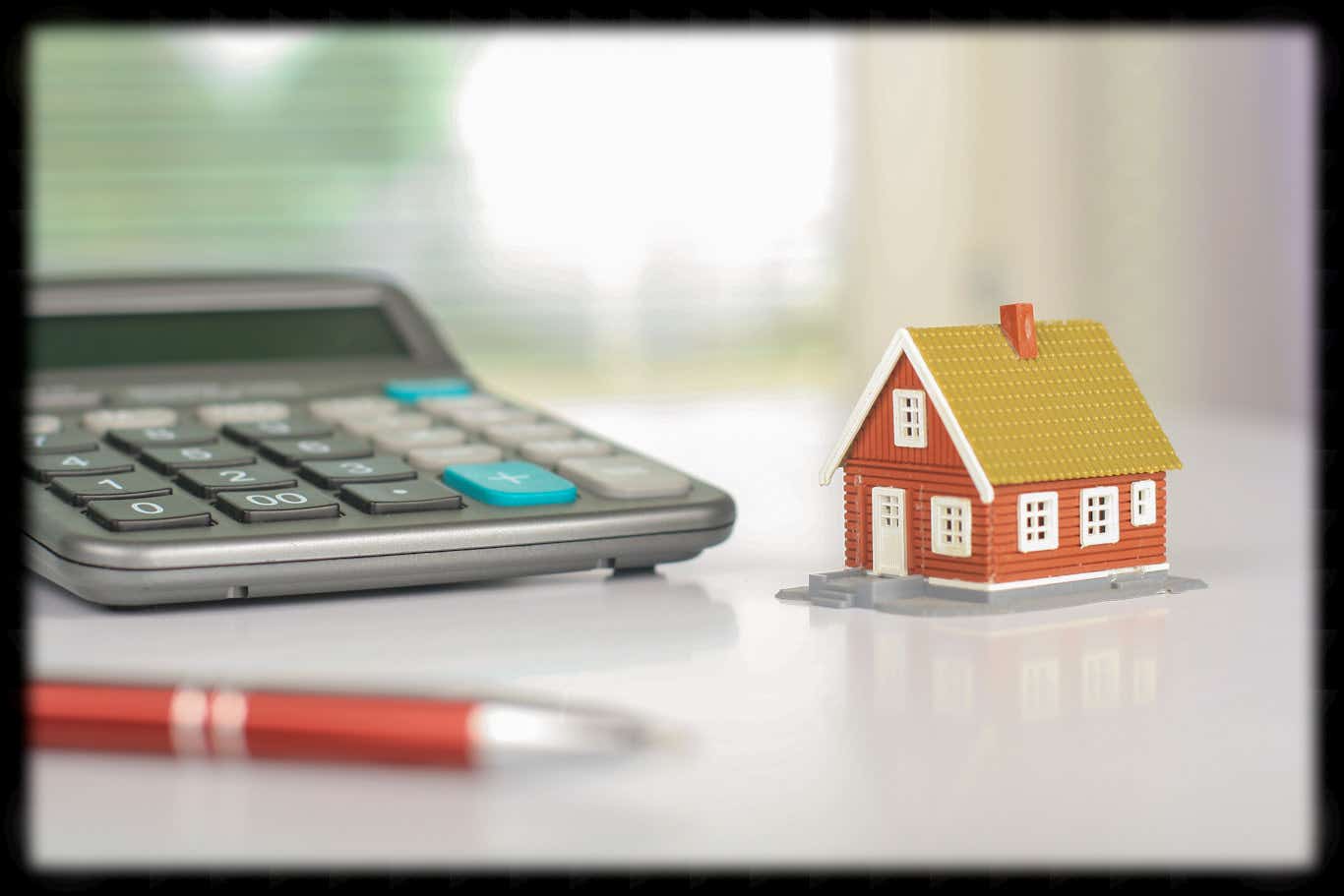 A large calculator sits on a shiny desk next to a model of a small red house. There are blurred vertical blinds at a window in the background and a blurred red pen in the foreground.