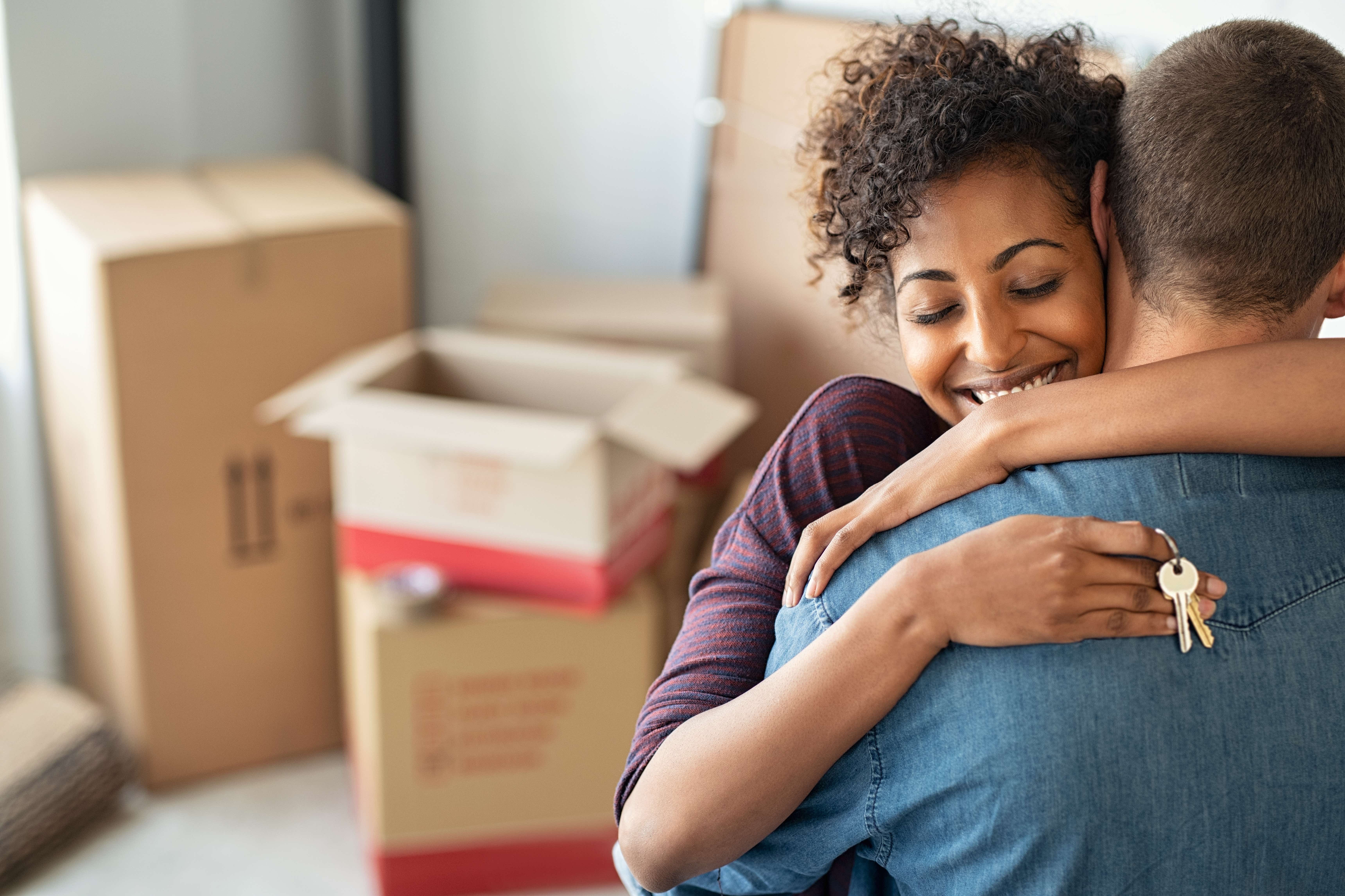 A couple embrace, an Asian lady faces toward the camera with a big smile and holding a set of keys, her partner faces away in her arms. In the background are a selection of out of focus cardboard boxes