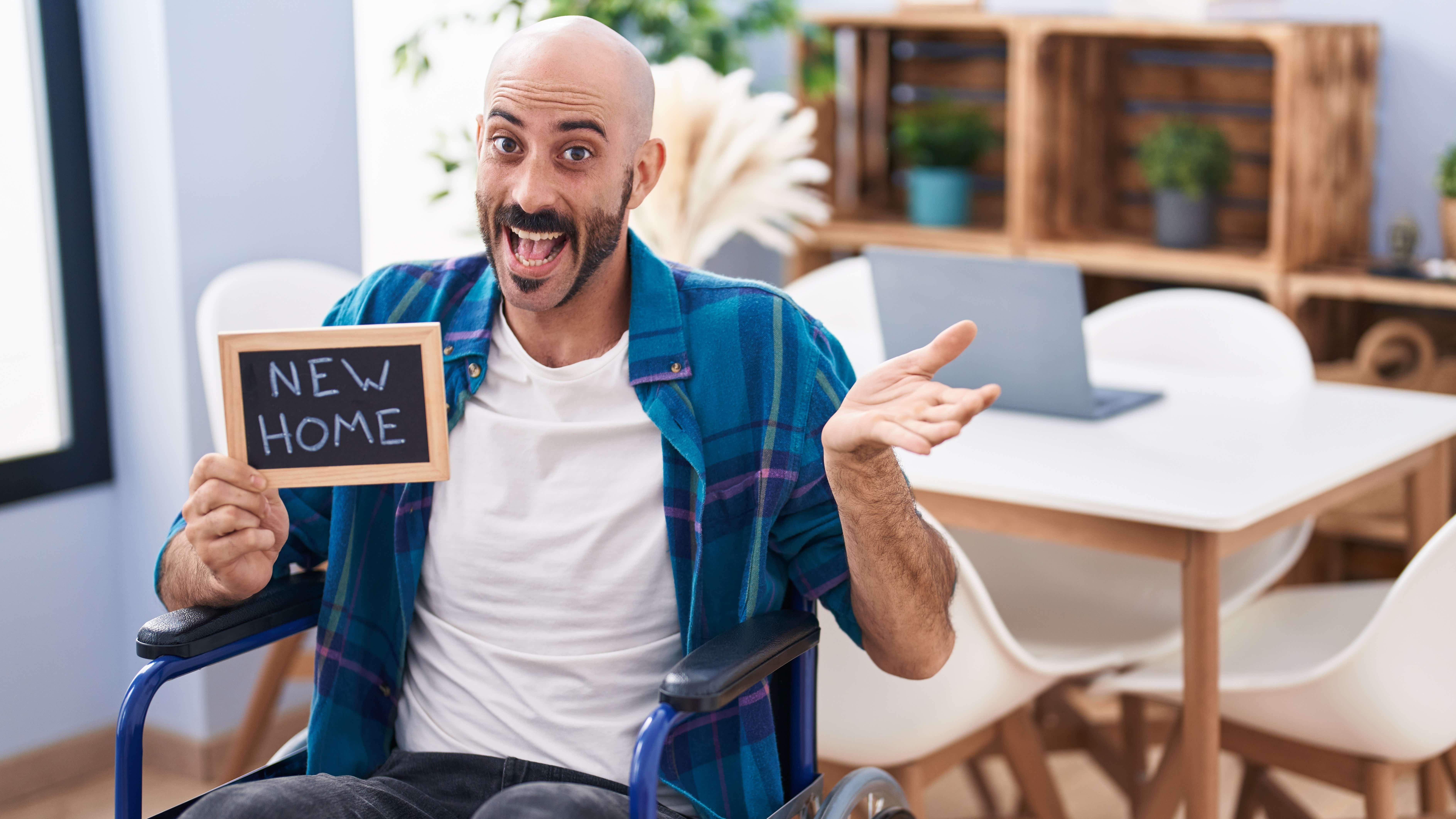 A young man in a wheelchair sits in a lounge holding a sign that has 'new home' written in chalk. He is surrounded by furniture and is smiling widely.