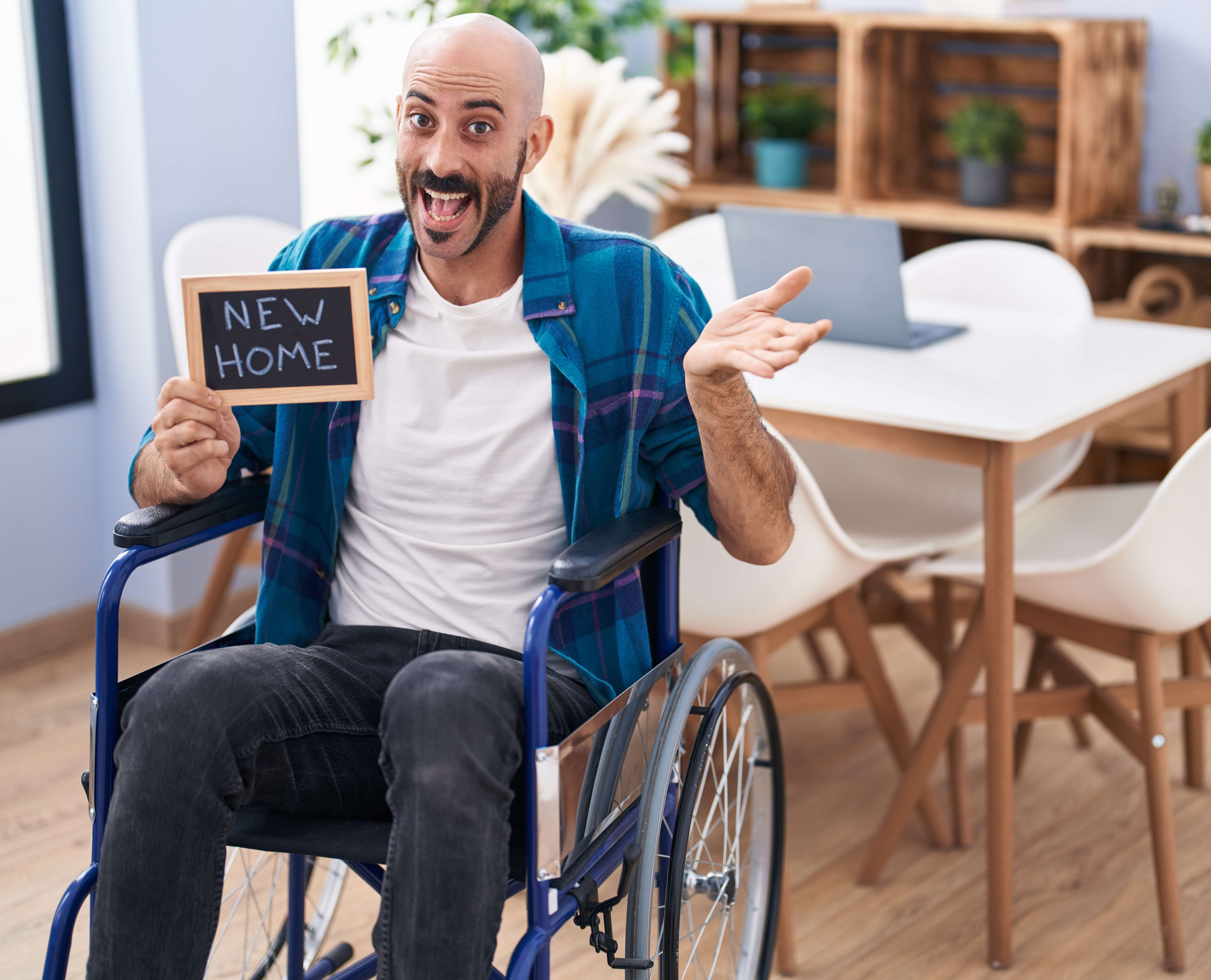 A young man in a wheelchair sits in a lounge holding a sign that has 'new home' written in chalk. He is surrounded by furniture and is smiling widely.