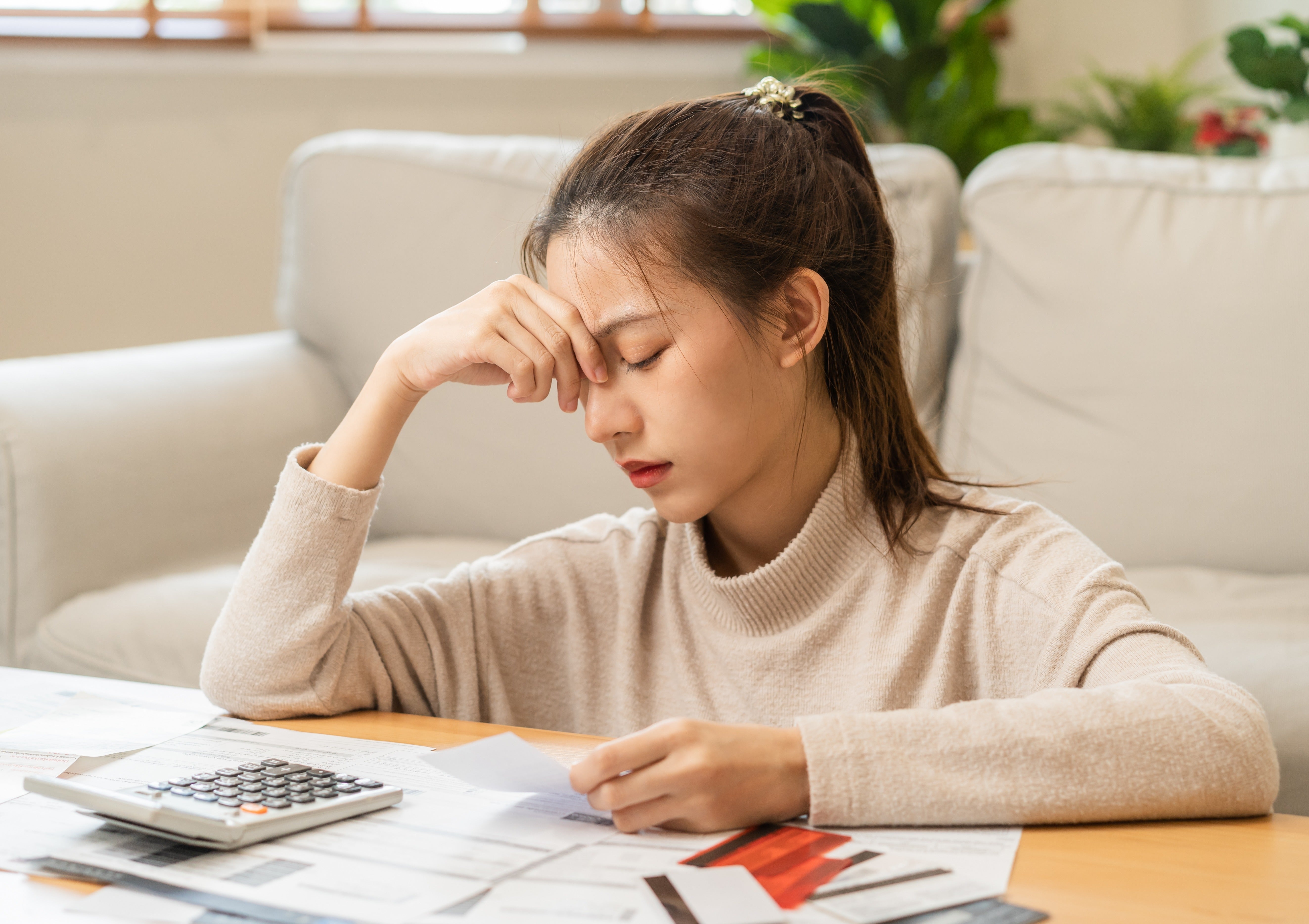 A young Asian woman is sitting on the floor next to a coffee table covered with bills. She has a look of deep concern, a furrowed brow and her hand is covering one of her eyes. 