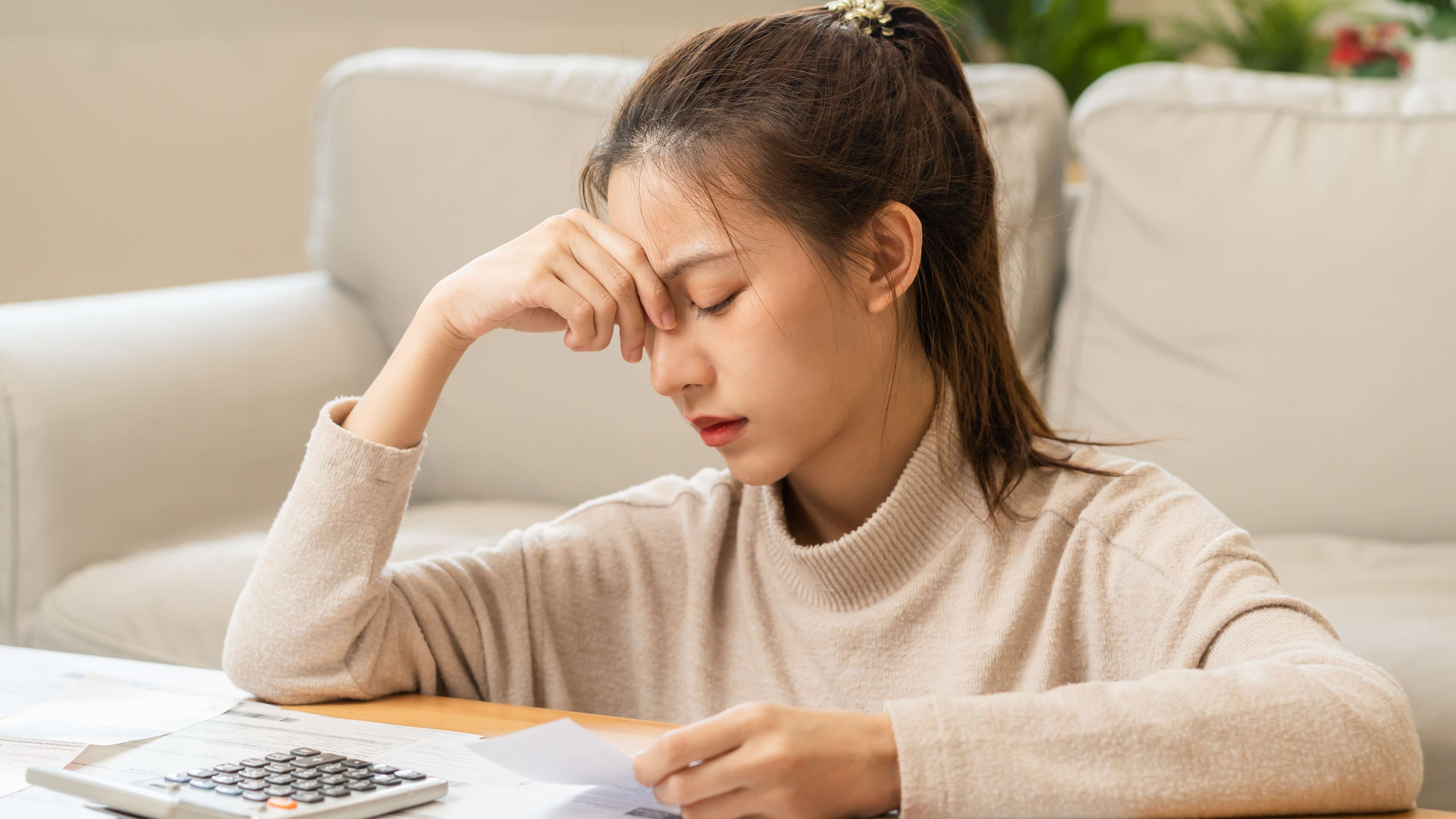 A young Asian woman is sitting on the floor next to a coffee table covered with bills. She has a look of deep concern, a furrowed brow and her hand is covering one of her eyes.