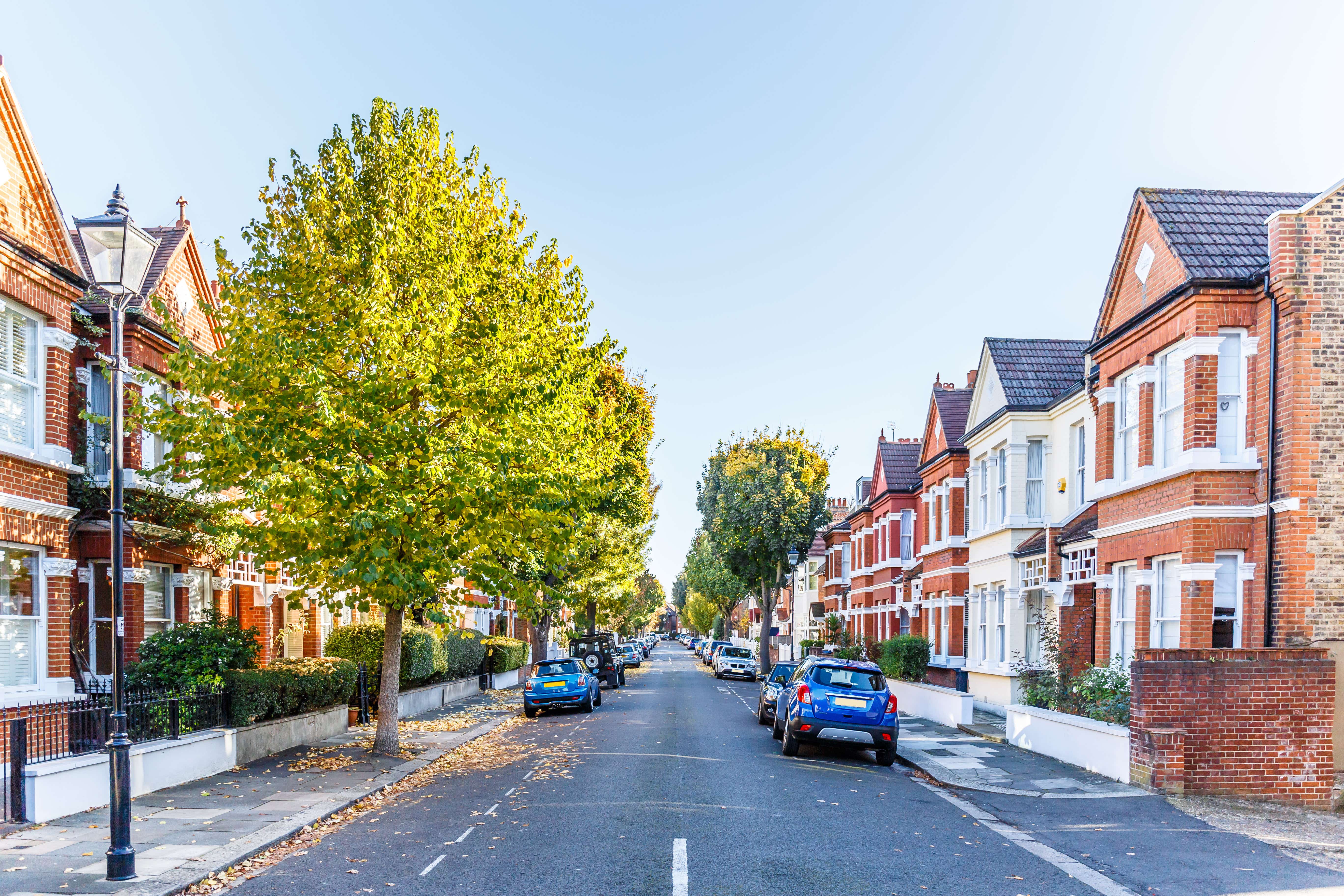 A tree lined street with mid-sized houses on either side of the road and cars parked along the kerbs.