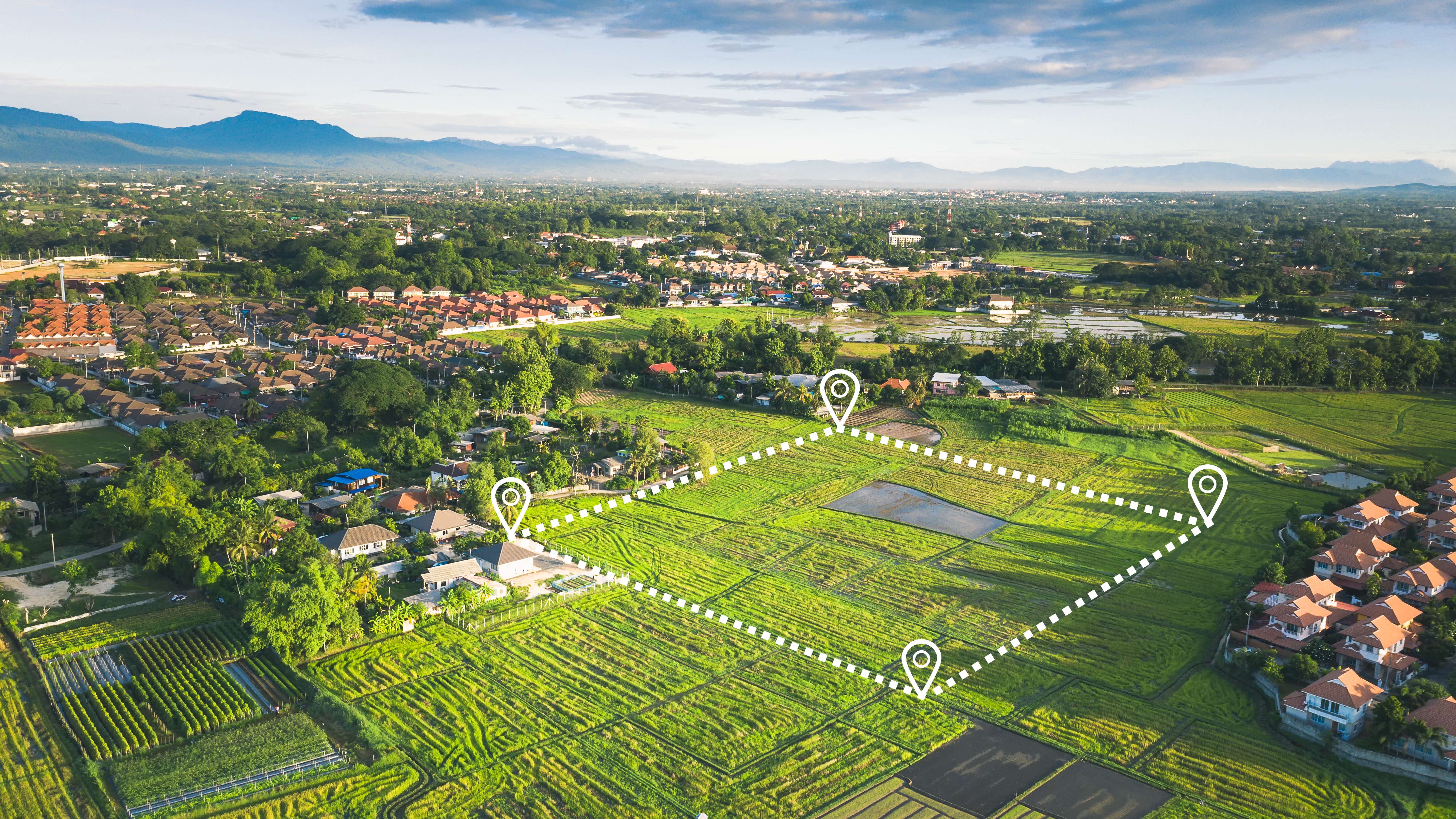 A n aerial photo shows semi-rural land from above, with a number of grassy fields and some buildings visible in the distance. There is a white dotted line in the shape of a square demarking a plot of land for sale