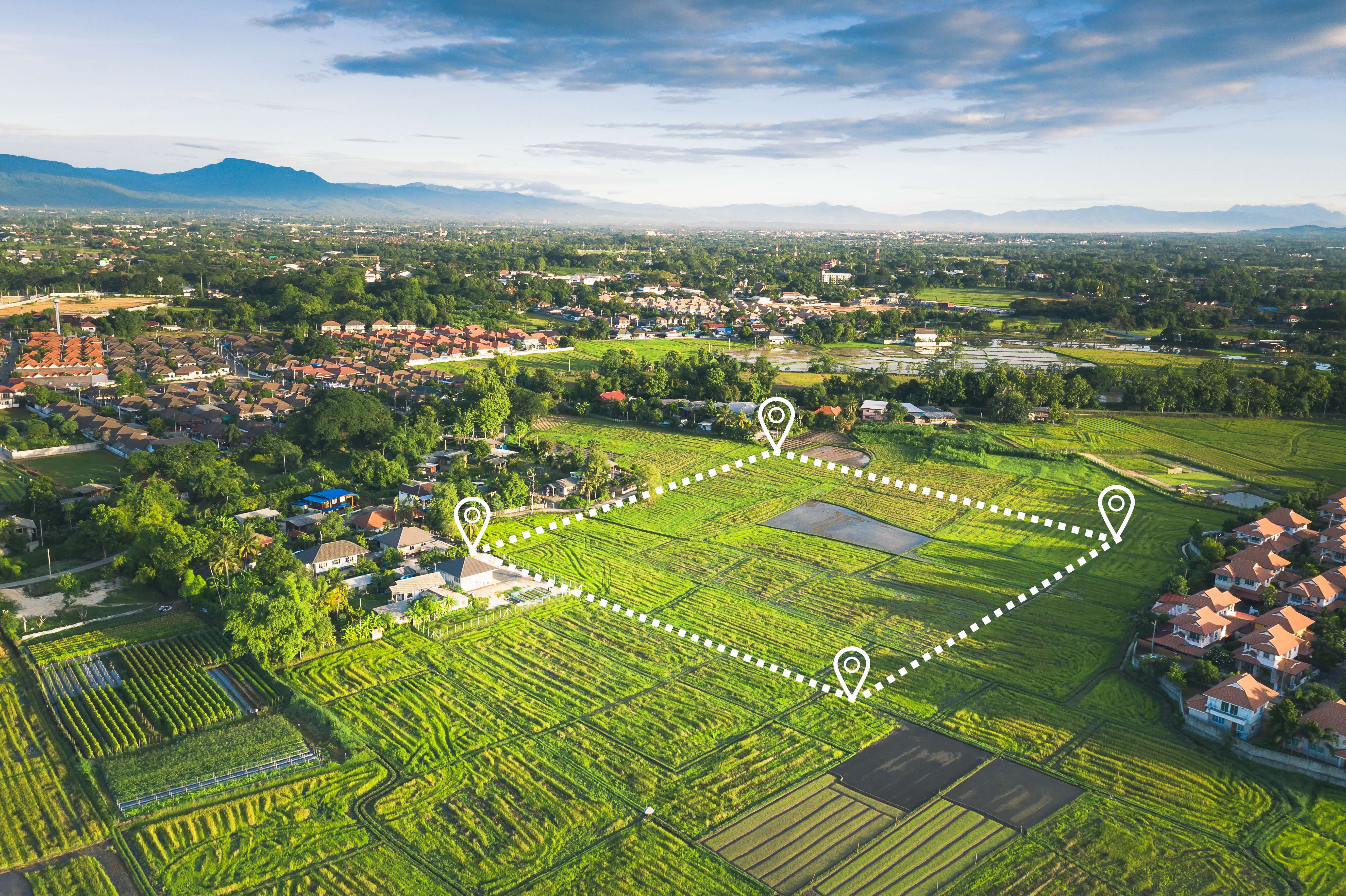 A n aerial photo shows semi-rural land from above, with a number of grassy fields and some buildings visible in the distance. There is a white dotted line in the shape of a square demarking a plot of land for sale