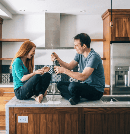 A redhead women and brunette man sit on their kitchen counter with an open bottle of champagne, celebrating their new home