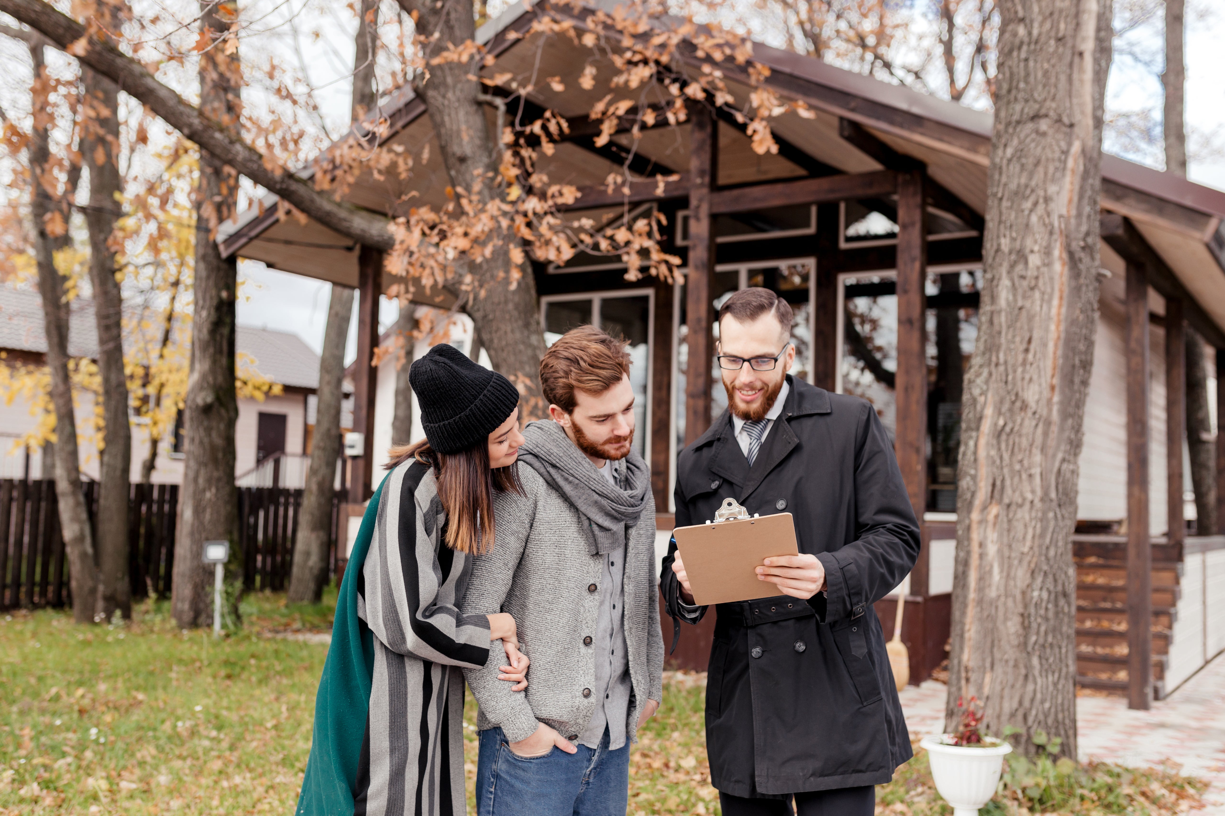 A young couple stand in front of a vacant property with a small areas of grass and some trees in the front garden. Another man is going through a checklist with them on a clipboard. All 3 are smiling