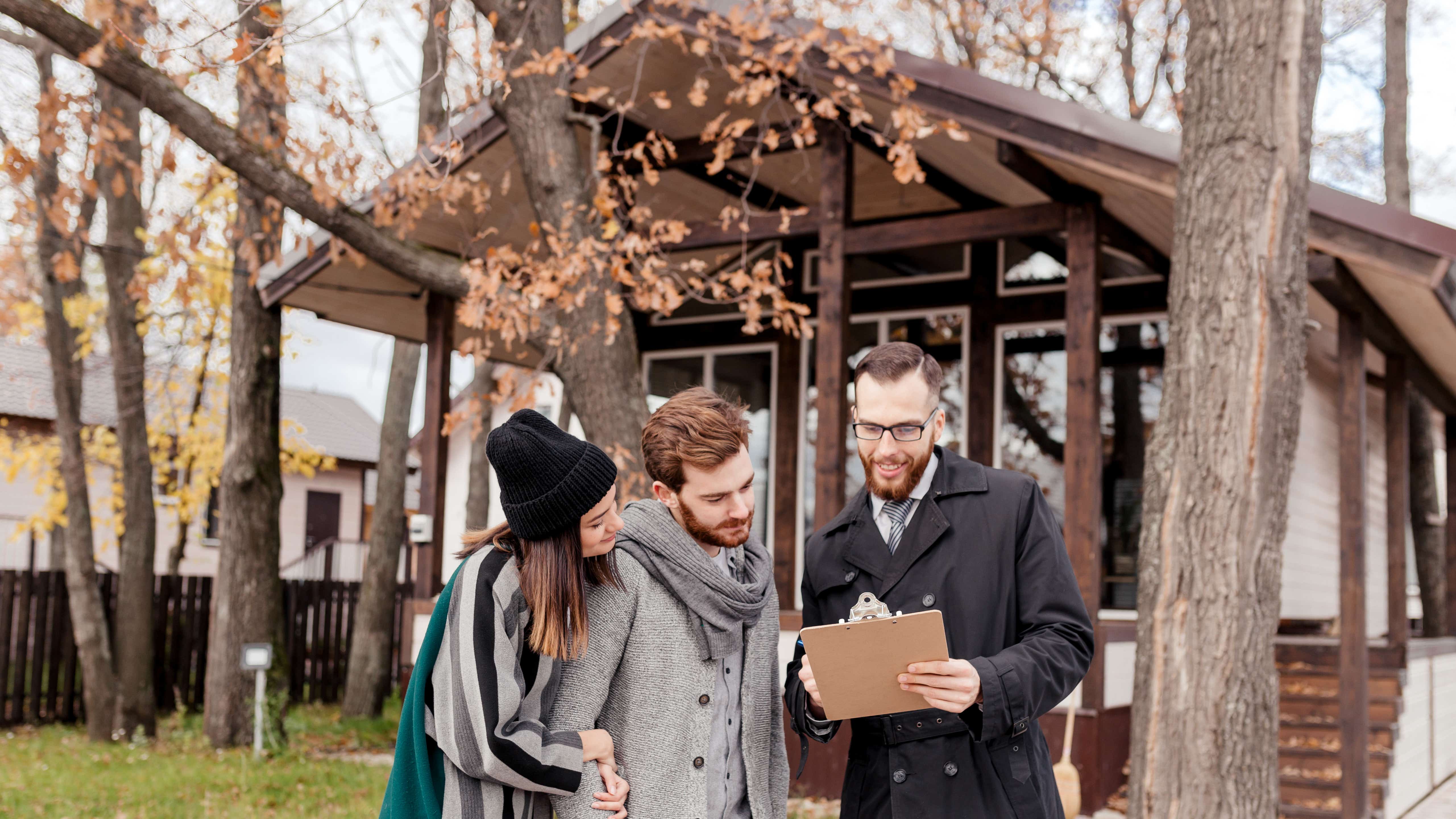 A young couple stand in front of a vacant property with a small areas of grass and some trees in the front garden. Another man is going through a checklist with them on a clipboard. All 3 are smiling