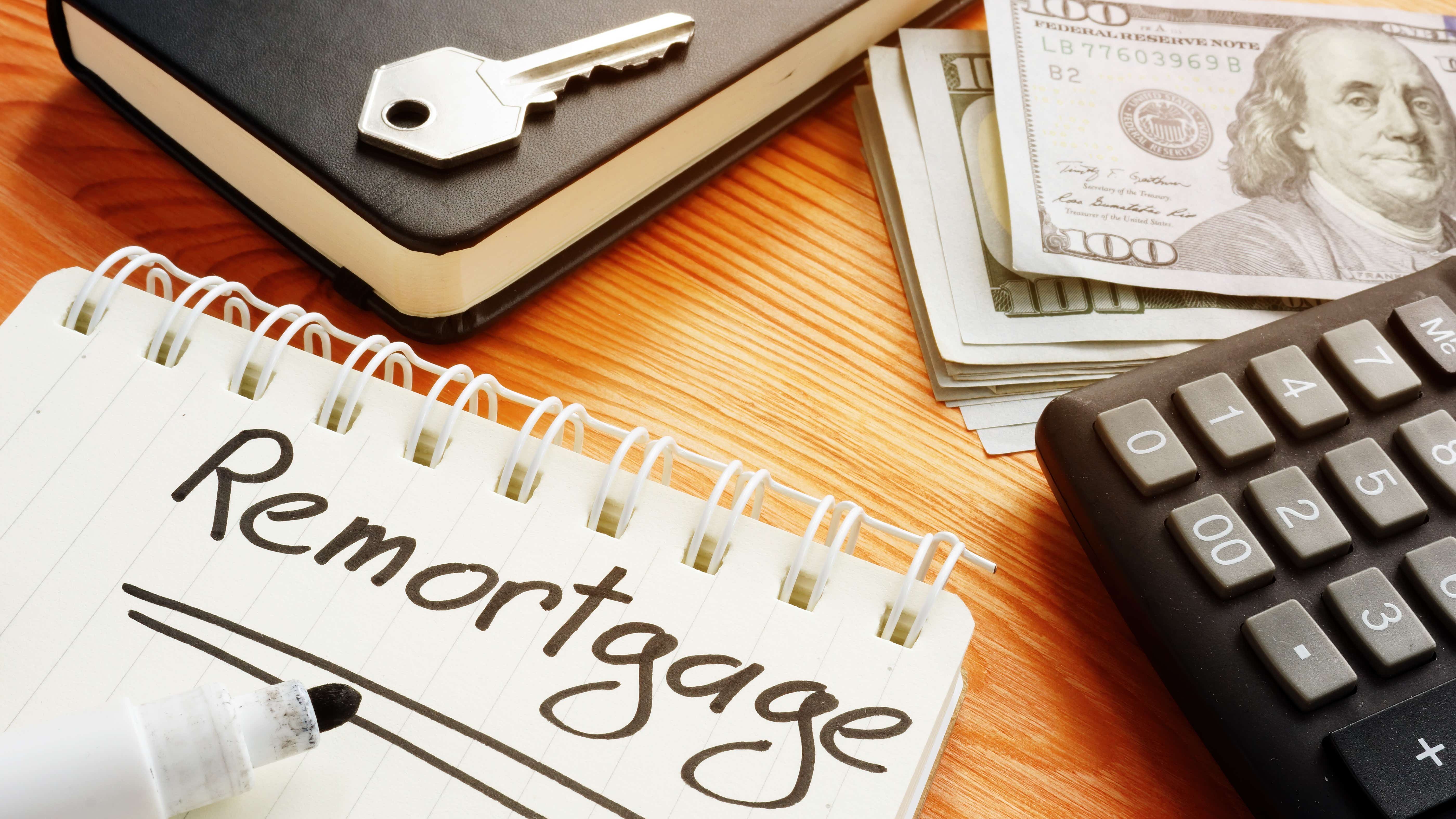 A wooden desk has the following items strewn on it: a calculator, some bank notes, a key, a diary and a notepad with the word 'Remortgage' hand written in bold black ink