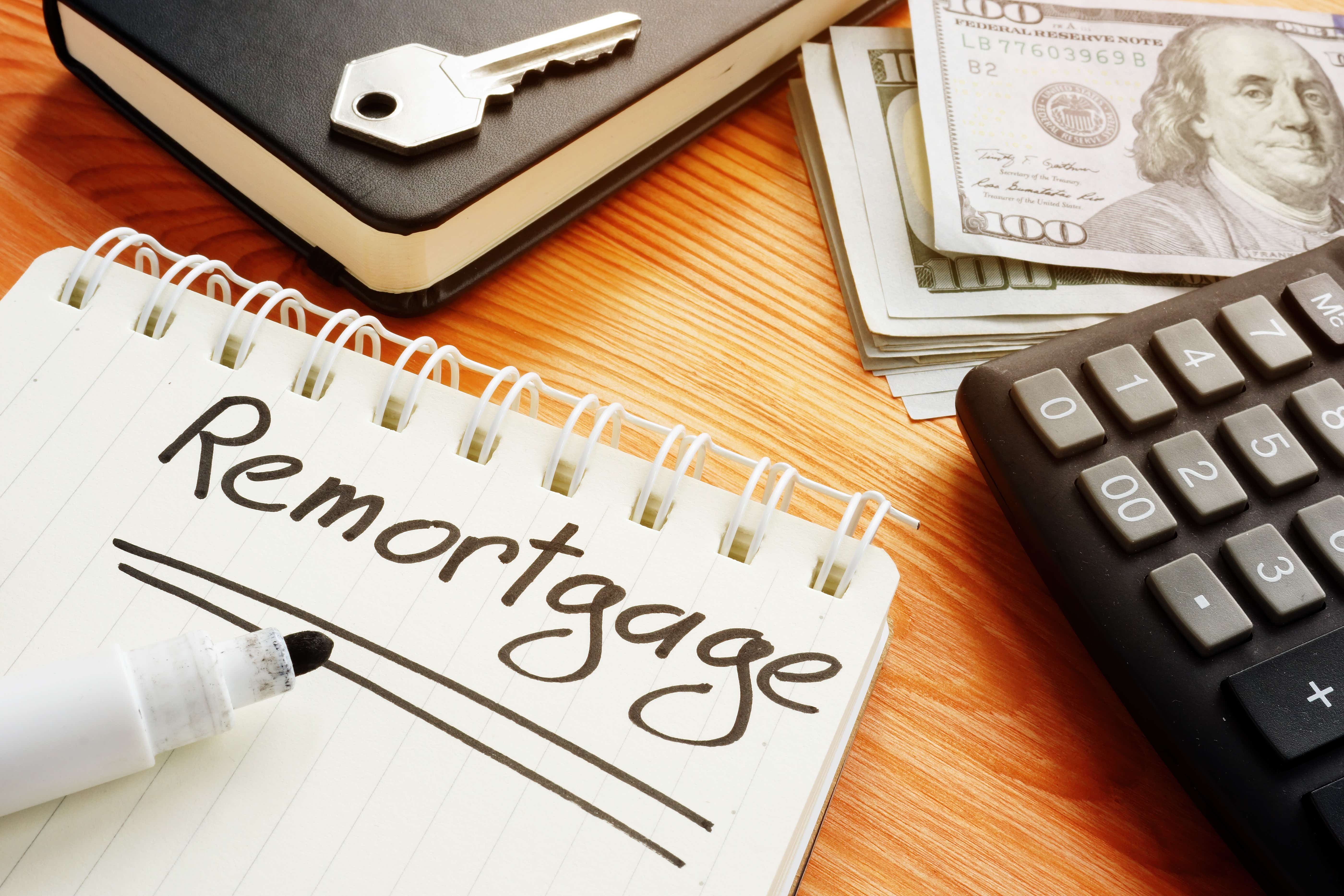 A wooden desk has the following items strewn on it: a calculator, some bank notes, a key, a diary and a notepad with the word 'Remortgage' hand written in bold black ink