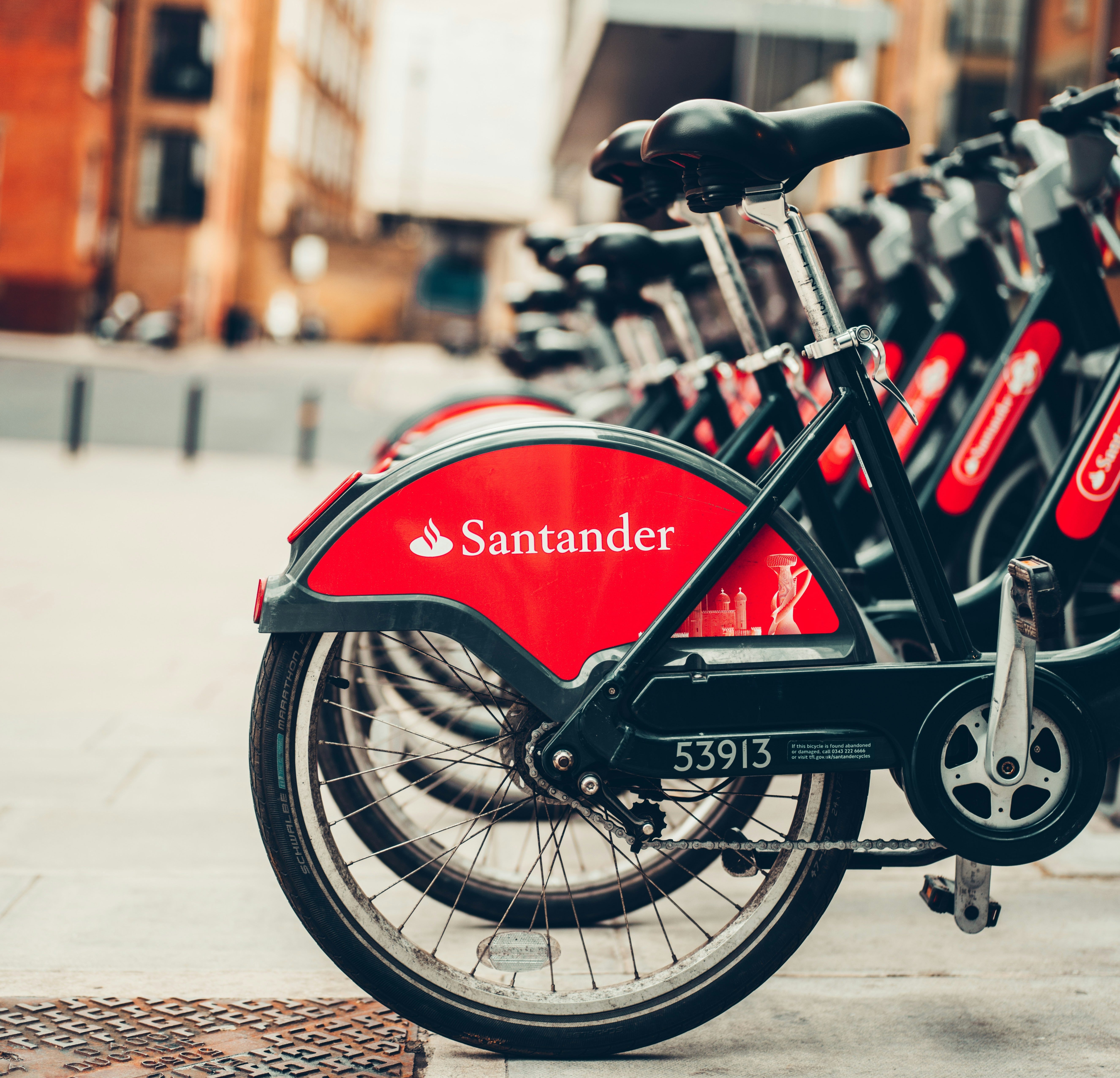 A row of bicycles are stationary with a blurred out city as the background. Each wheel has a red Santander logo displayed across it.
