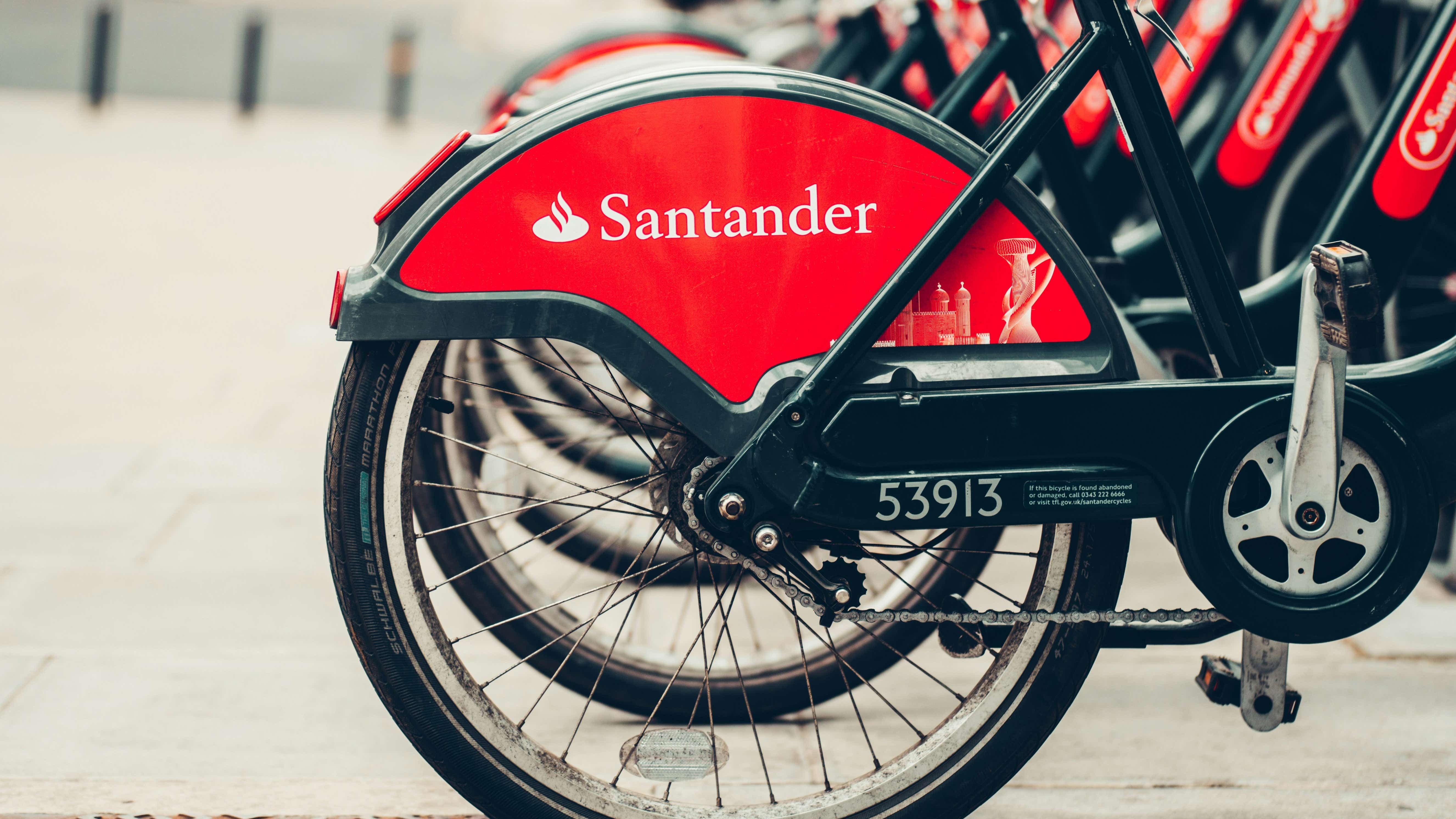 A row of bicycles are stationary with a blurred out city as the background. Each wheel has a red Santander logo displayed across it.