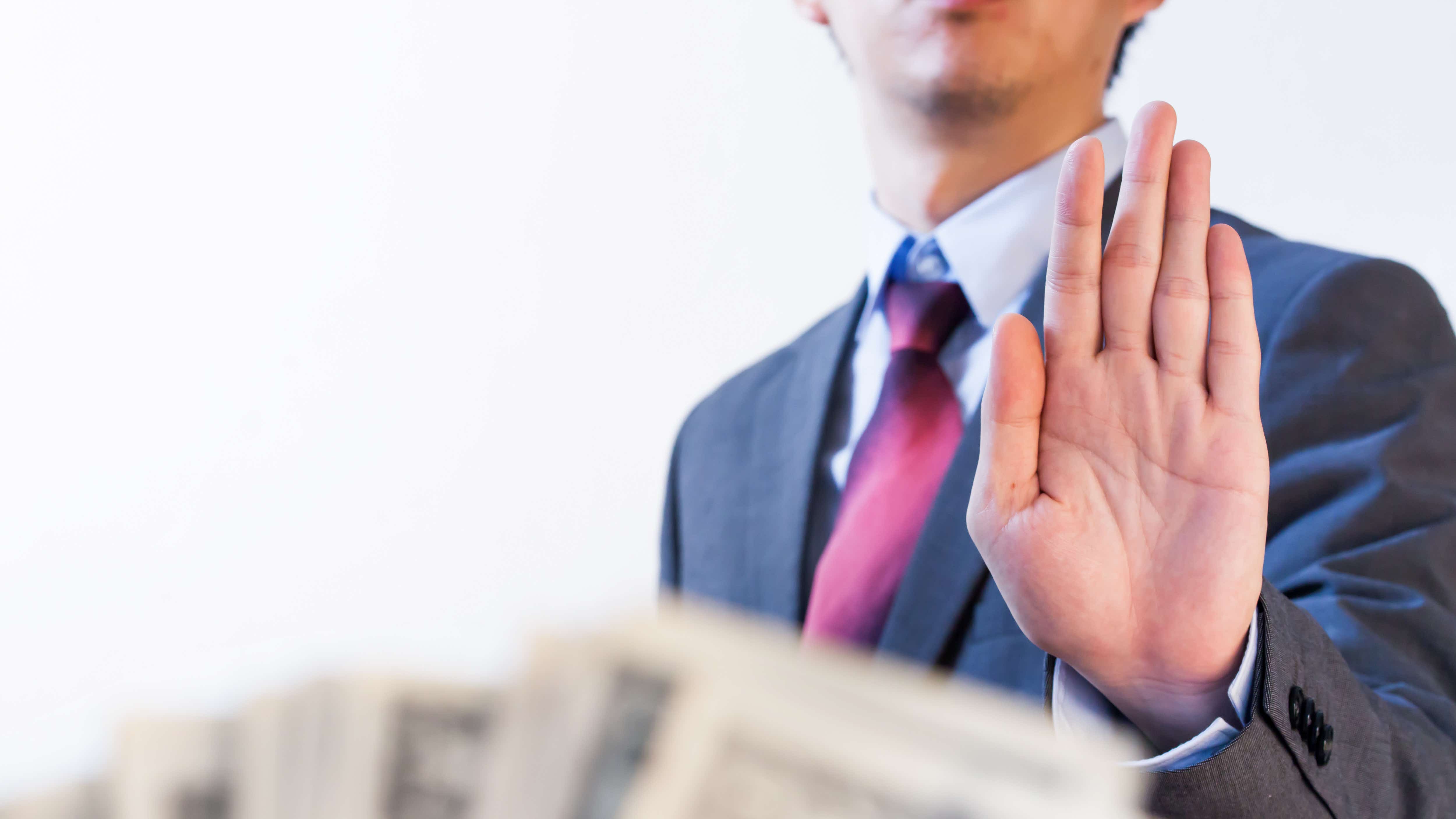 A person dressed in a suit has just their abdomen and hand visible. Their hand is raised up in a refusal position and in the foreground, someone else is offering up a fan of note currency.