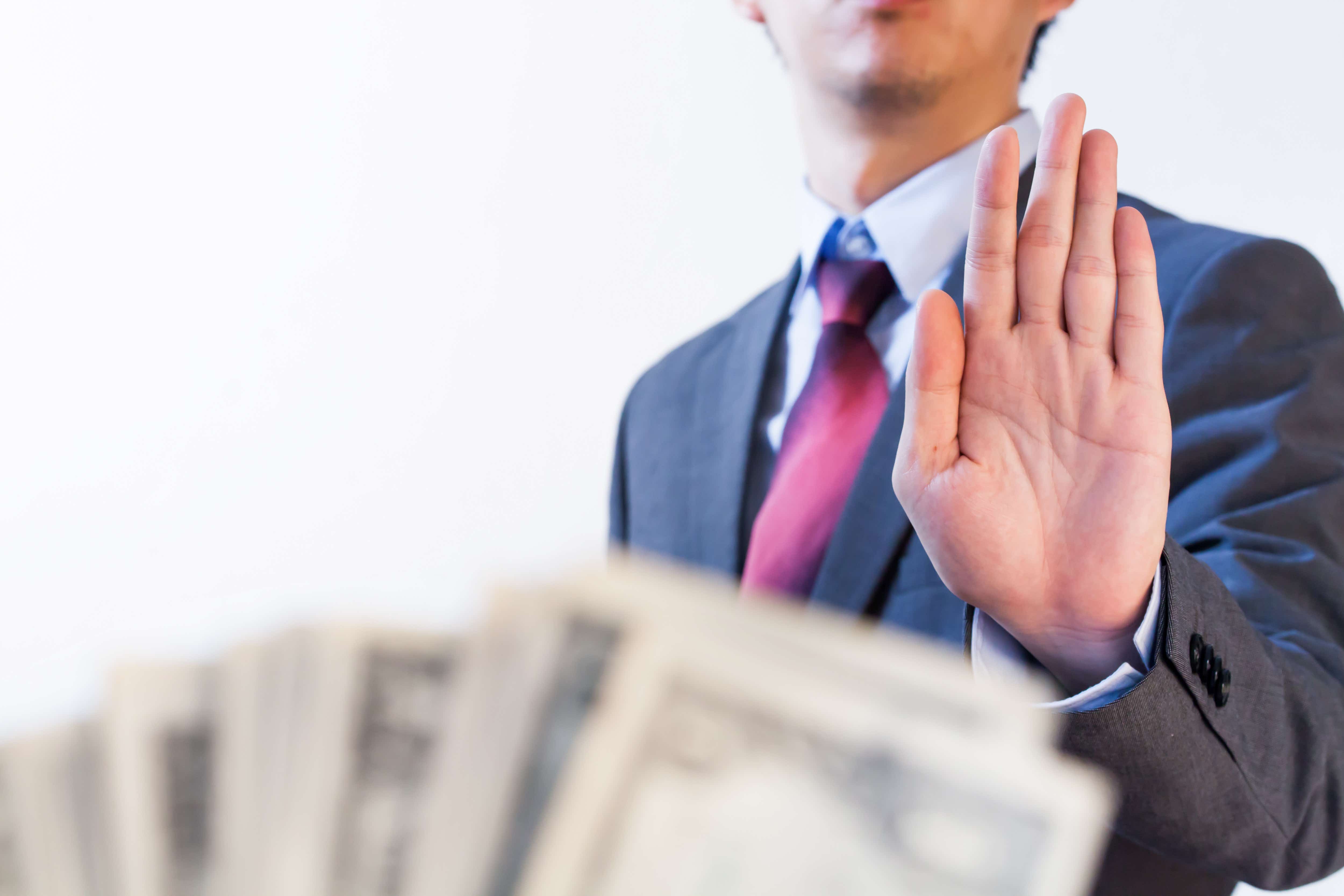 A person dressed in a suit has just their abdomen and hand visible. Their hand is raised up in a refusal position and in the foreground, someone else is offering up a fan of note currency.