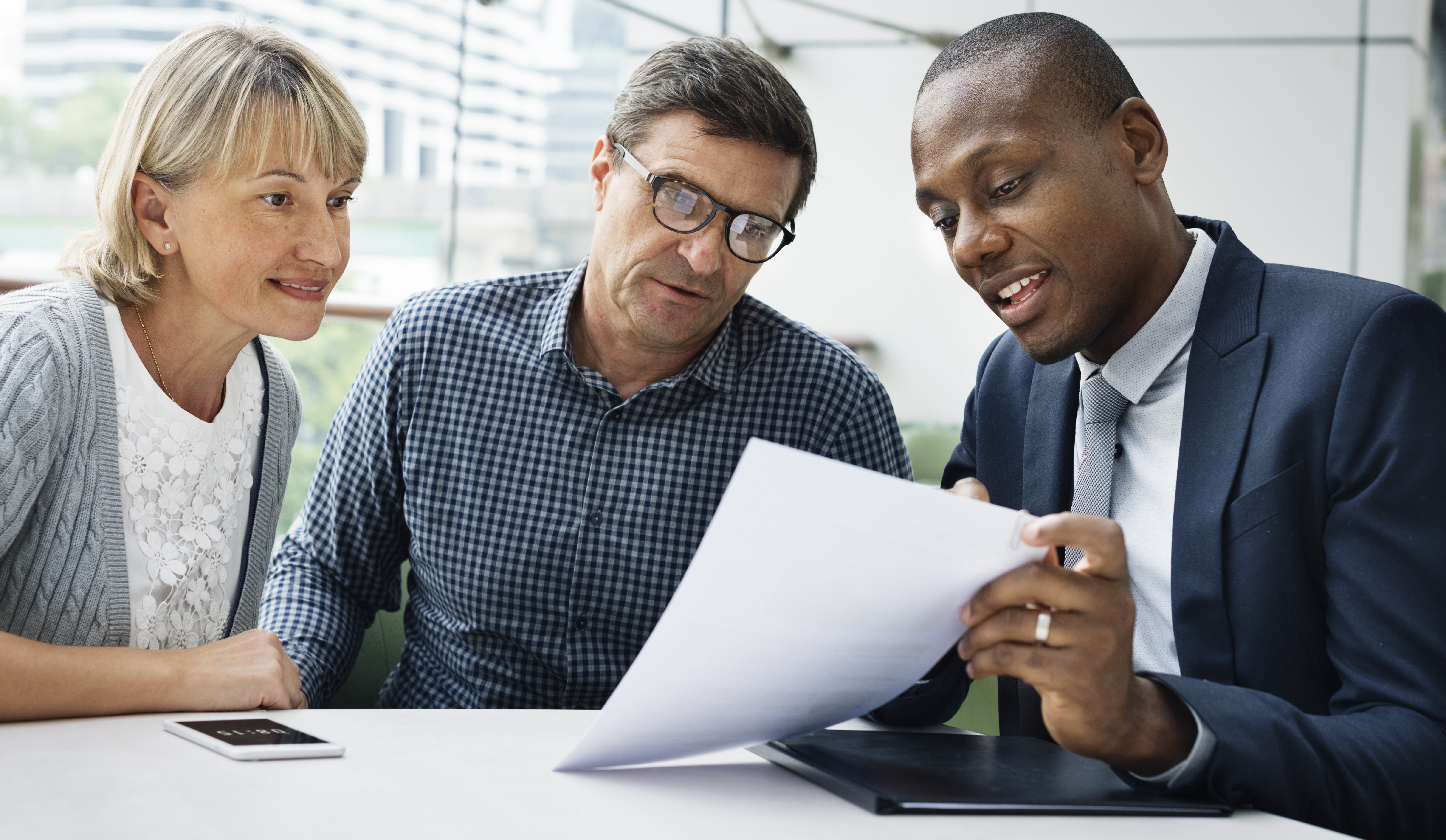 A black man in a suit and tie shows a middle-aged white couple some paperwork. They are sitting in front of large windows in an office, with a desk at the forefront