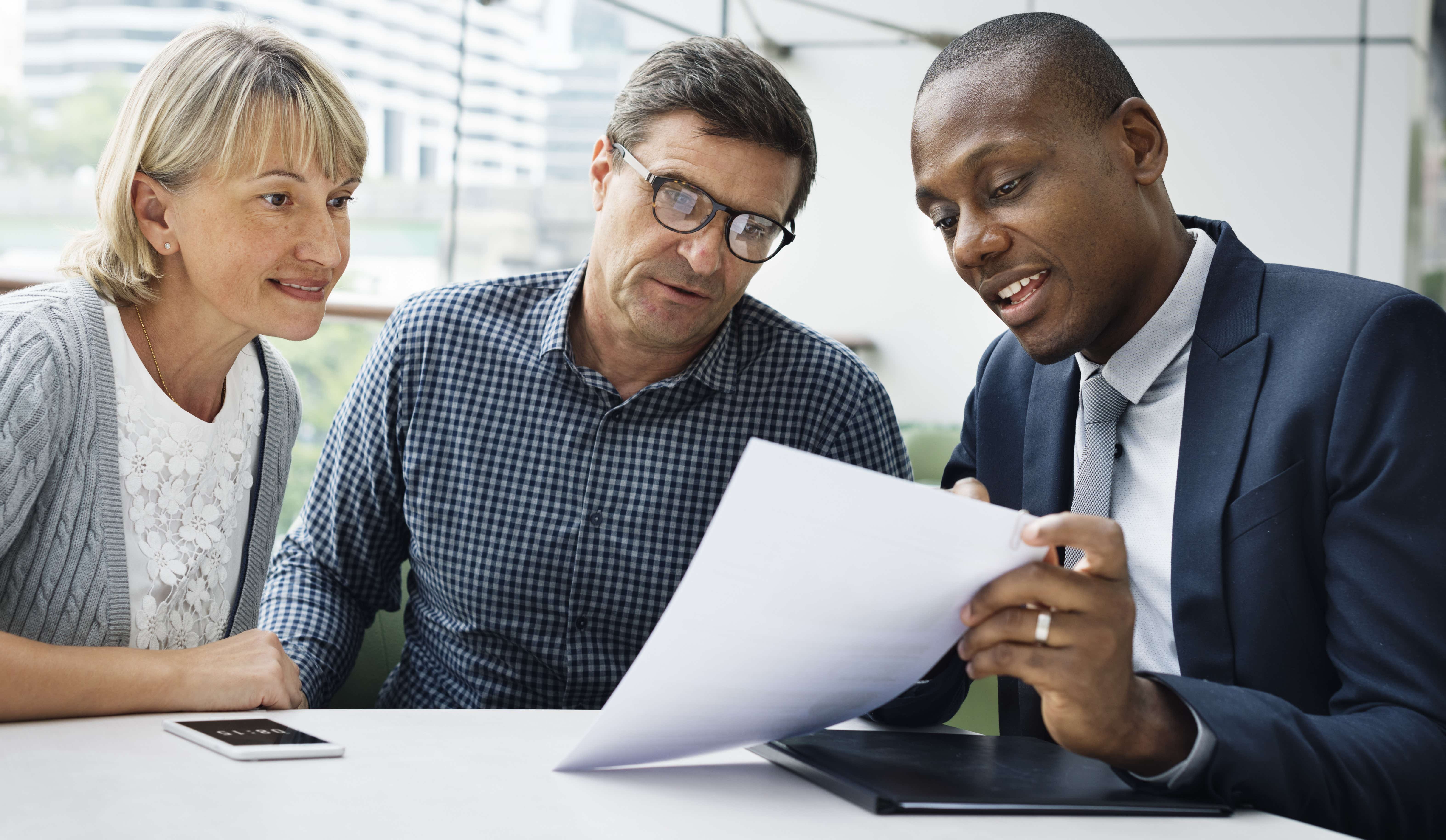 A black man in a suit and tie shows a middle-aged white couple some paperwork. They are sitting in front of large windows in an office, with a desk at the forefront