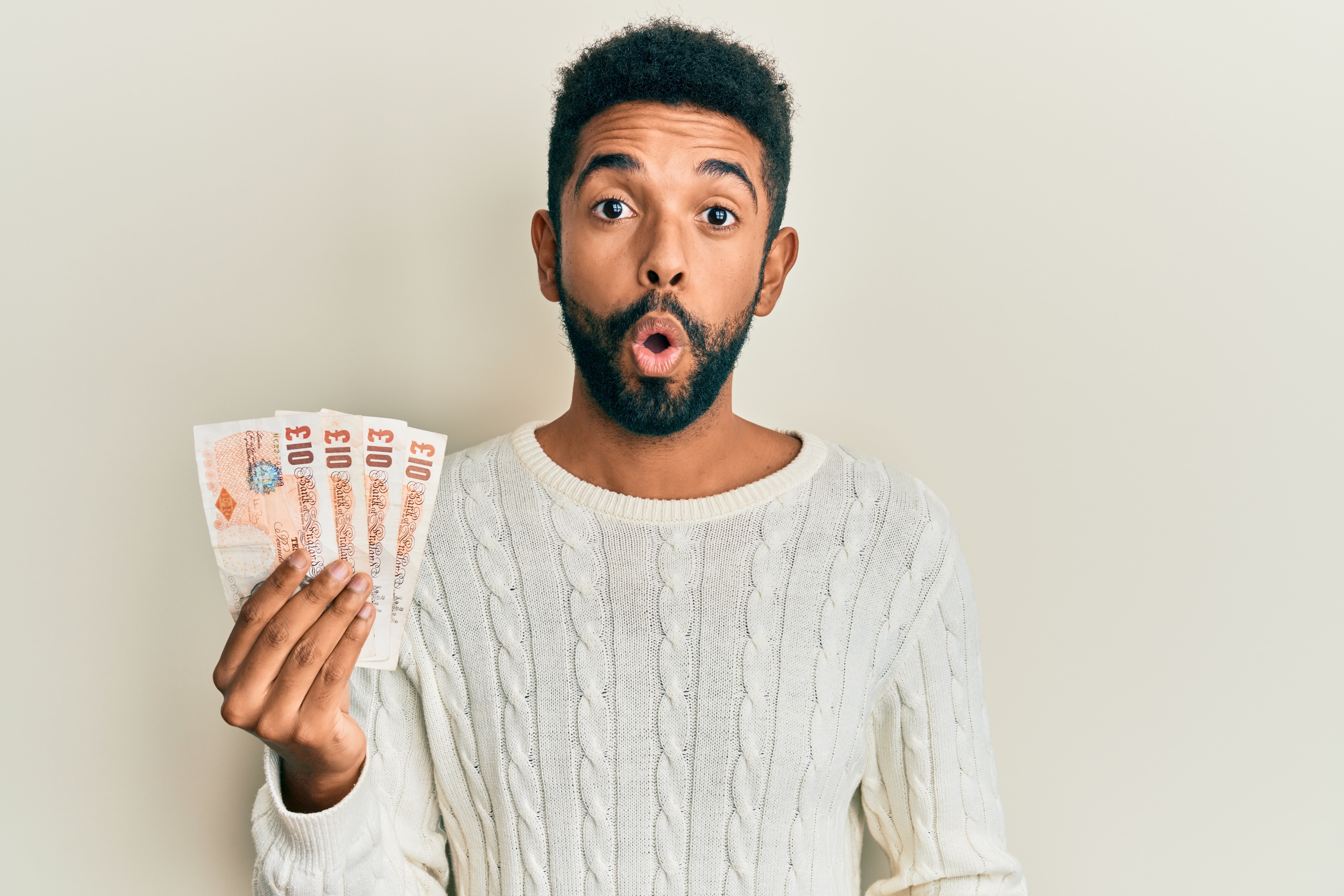 A young black male with a beard and a white knitted sweater holds up a fan of UK banknotes with a shocked and excited expression on his face