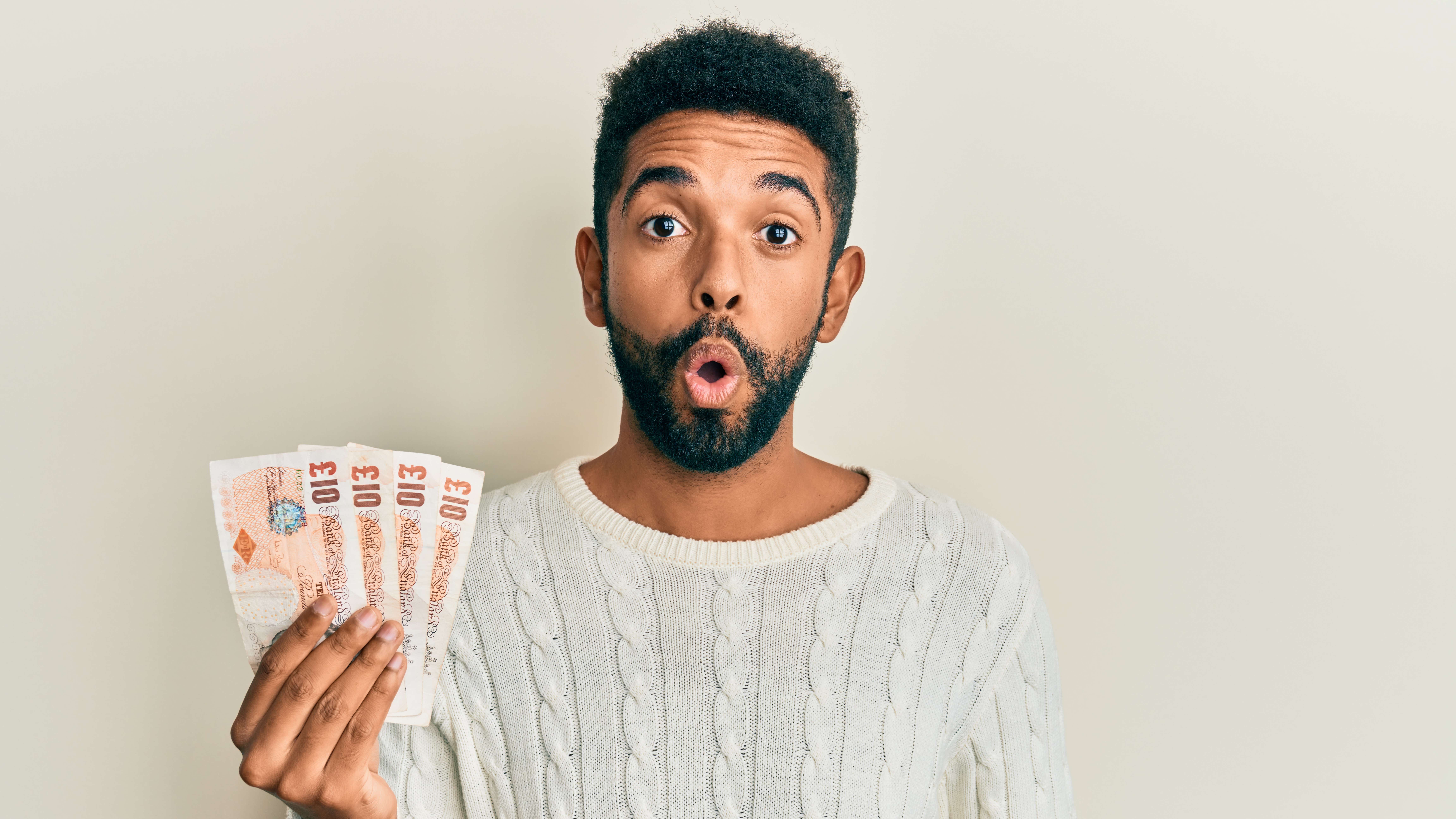 A young black male with a beard and a white knitted sweater holds up a fan of UK banknotes with a shocked and excited expression on his face