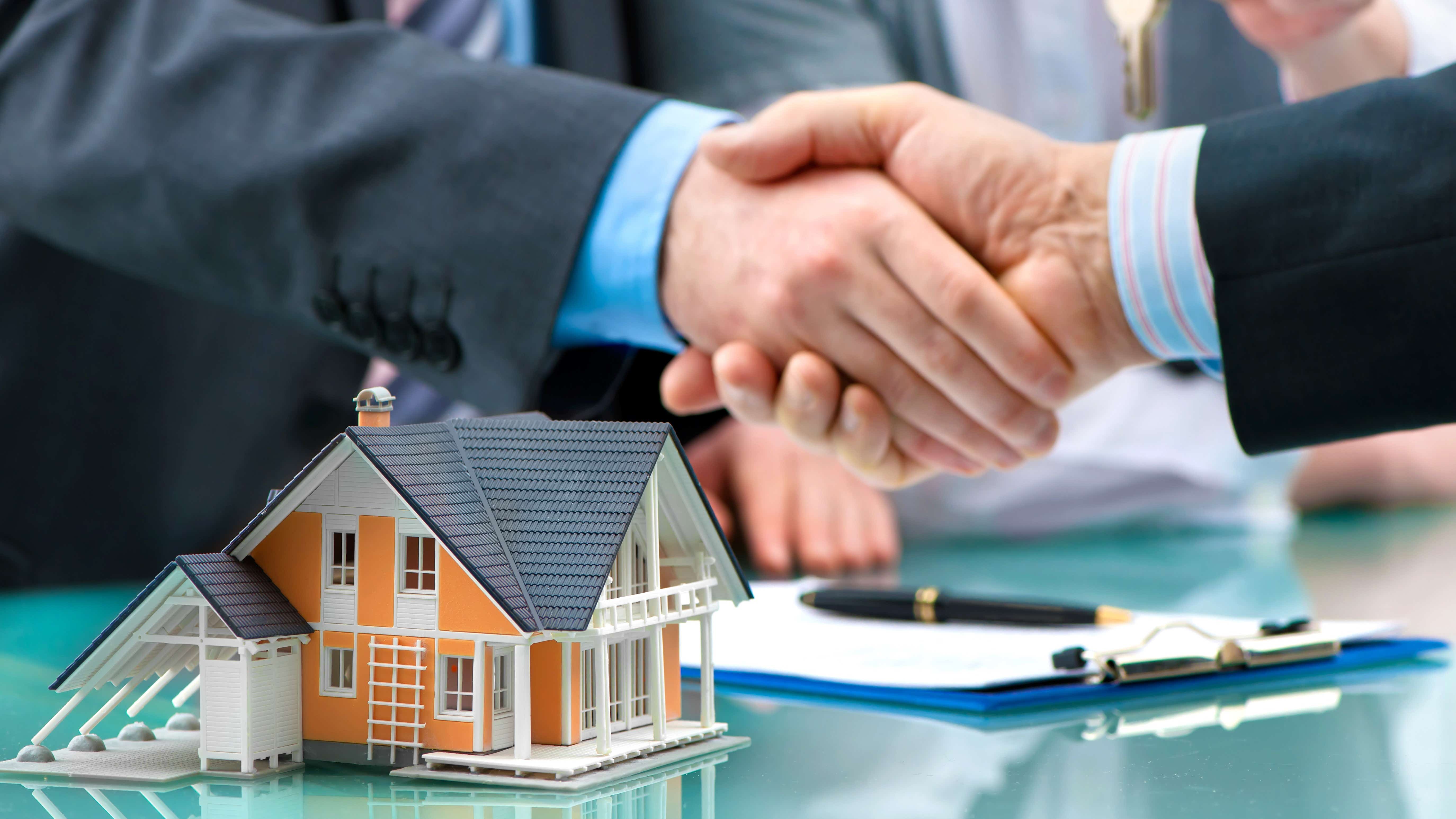Two hands are shaking across a table and in the background another hand holds a key. On the table is a tiny model of a house and a form with a pen