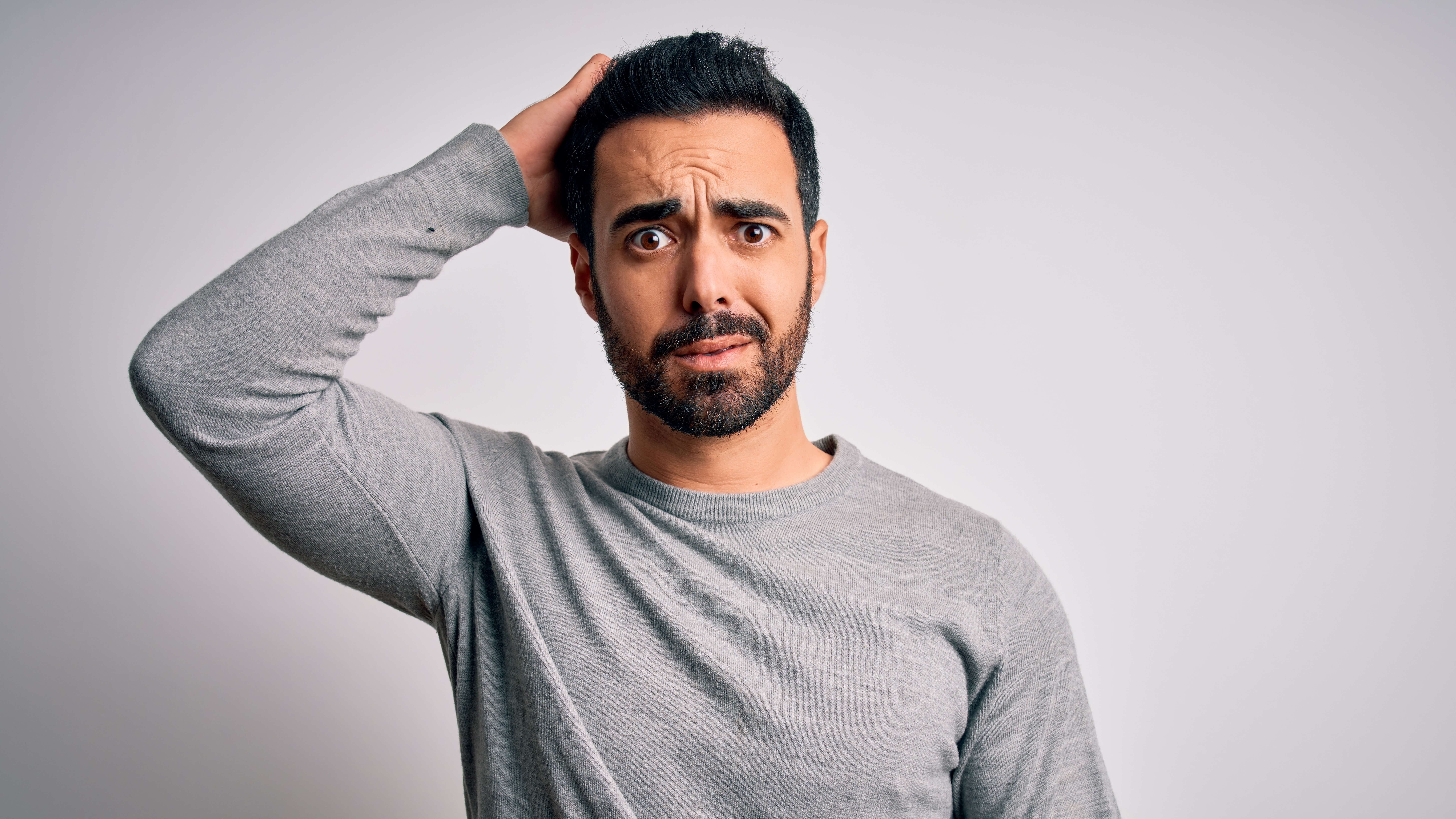 An olive-skinned man with black hair and beard and a grey sweater stands in front of a blank grey wall with his right arm raised behind his head and a look of deep confusion