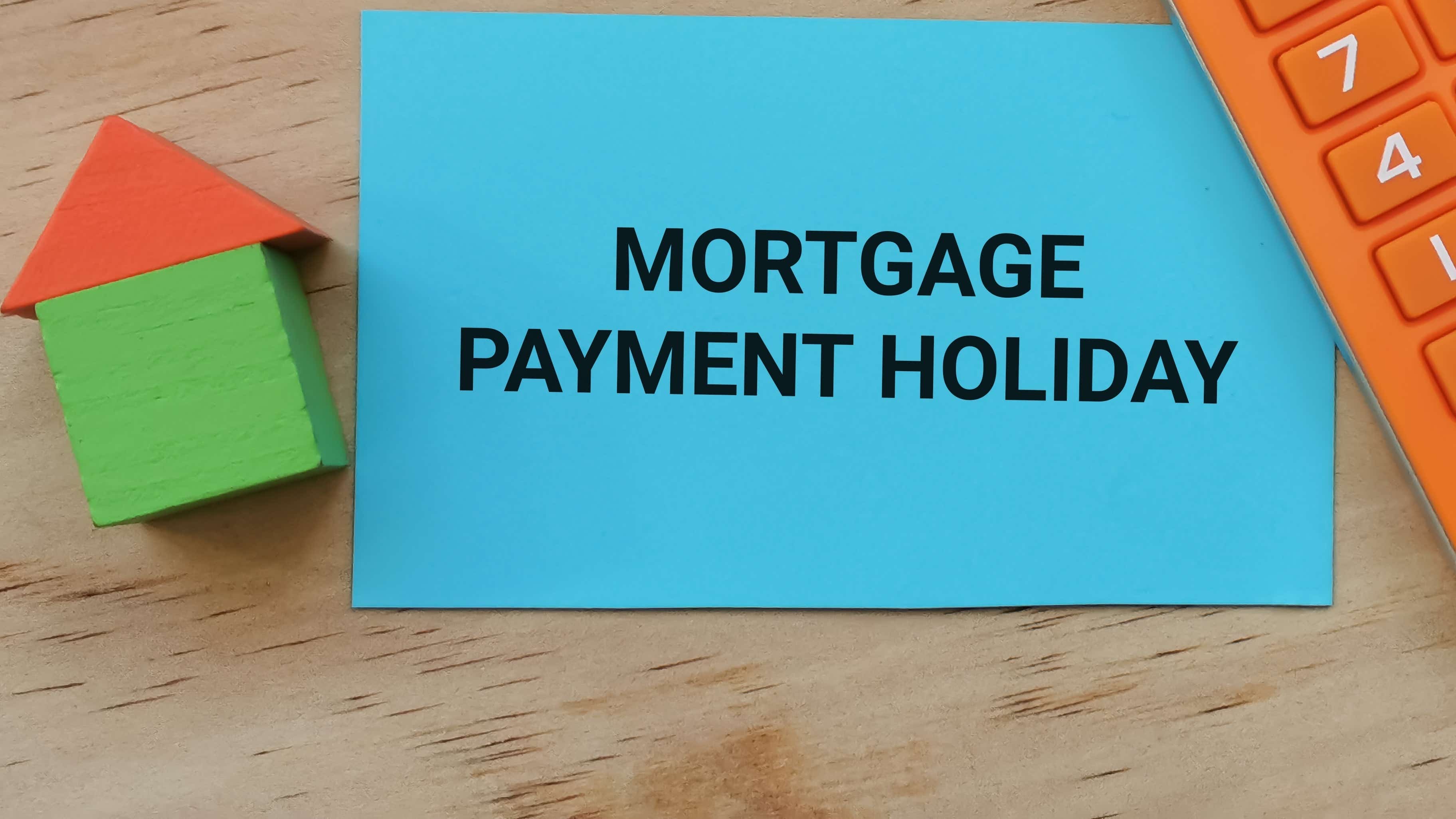 On a wooden desk lays an orange calculator, a small model of a green house with a red roof and a blue card with the words 'mortgage payment holiday' in bold print.