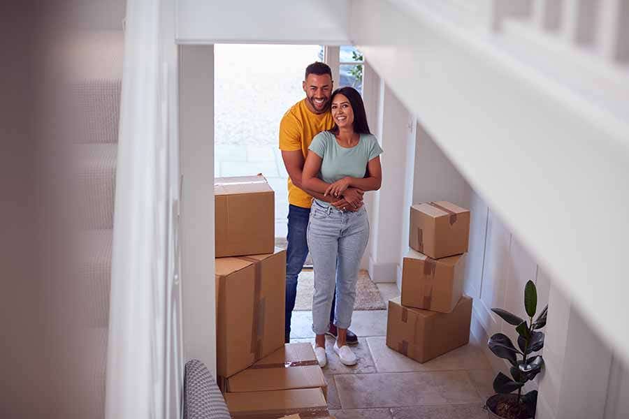 A male and female couple stand in the entrance hall of their new home with smiles on their faces. There is sun shining into the white hallway which is lined with cardboard boxes.