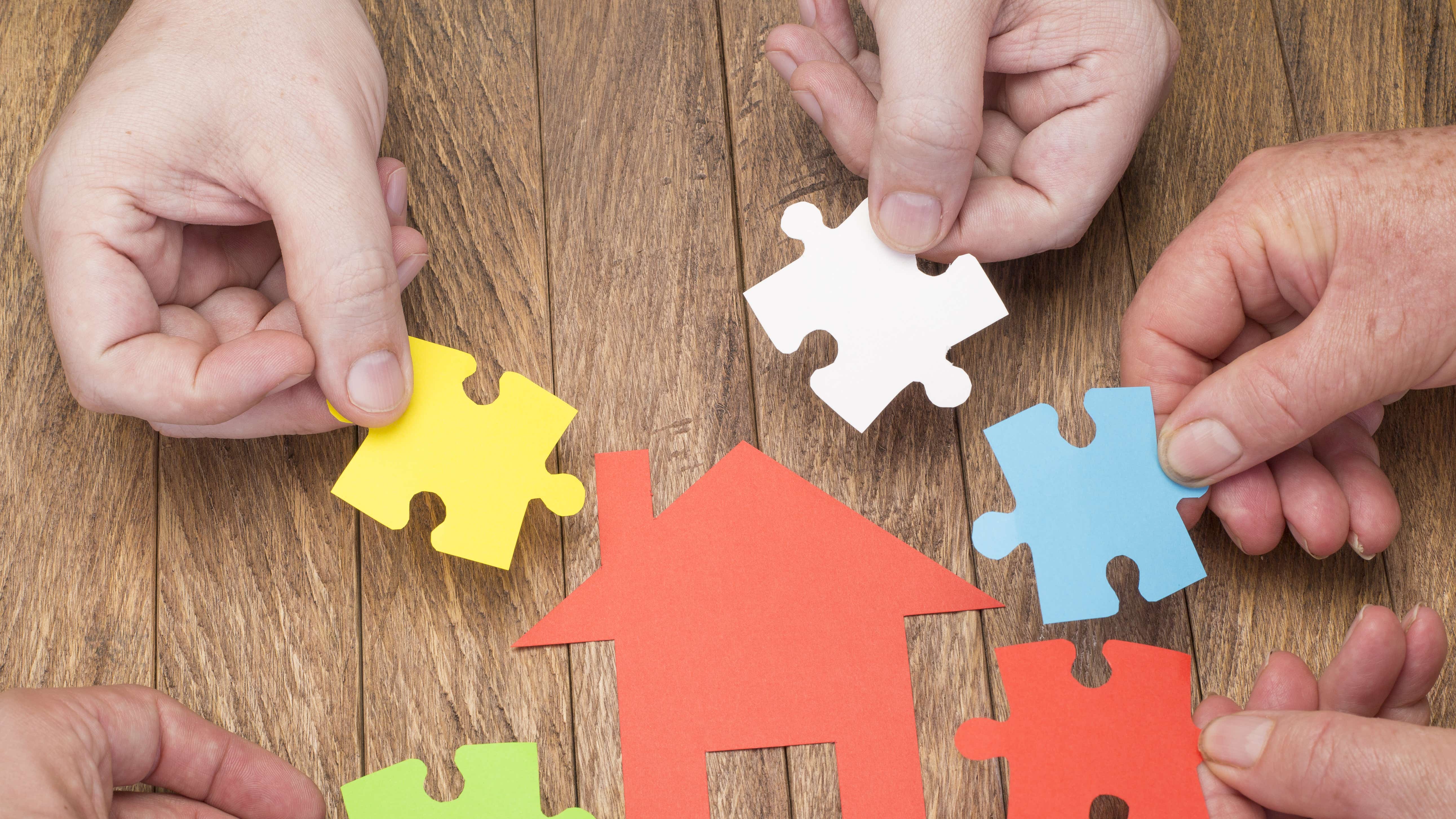 A red paper cut out of a house is laid onto a wooden desk. There are 4 hands from different people surrounding the house, each holding a paper cutout shaped like a jigsaw piece