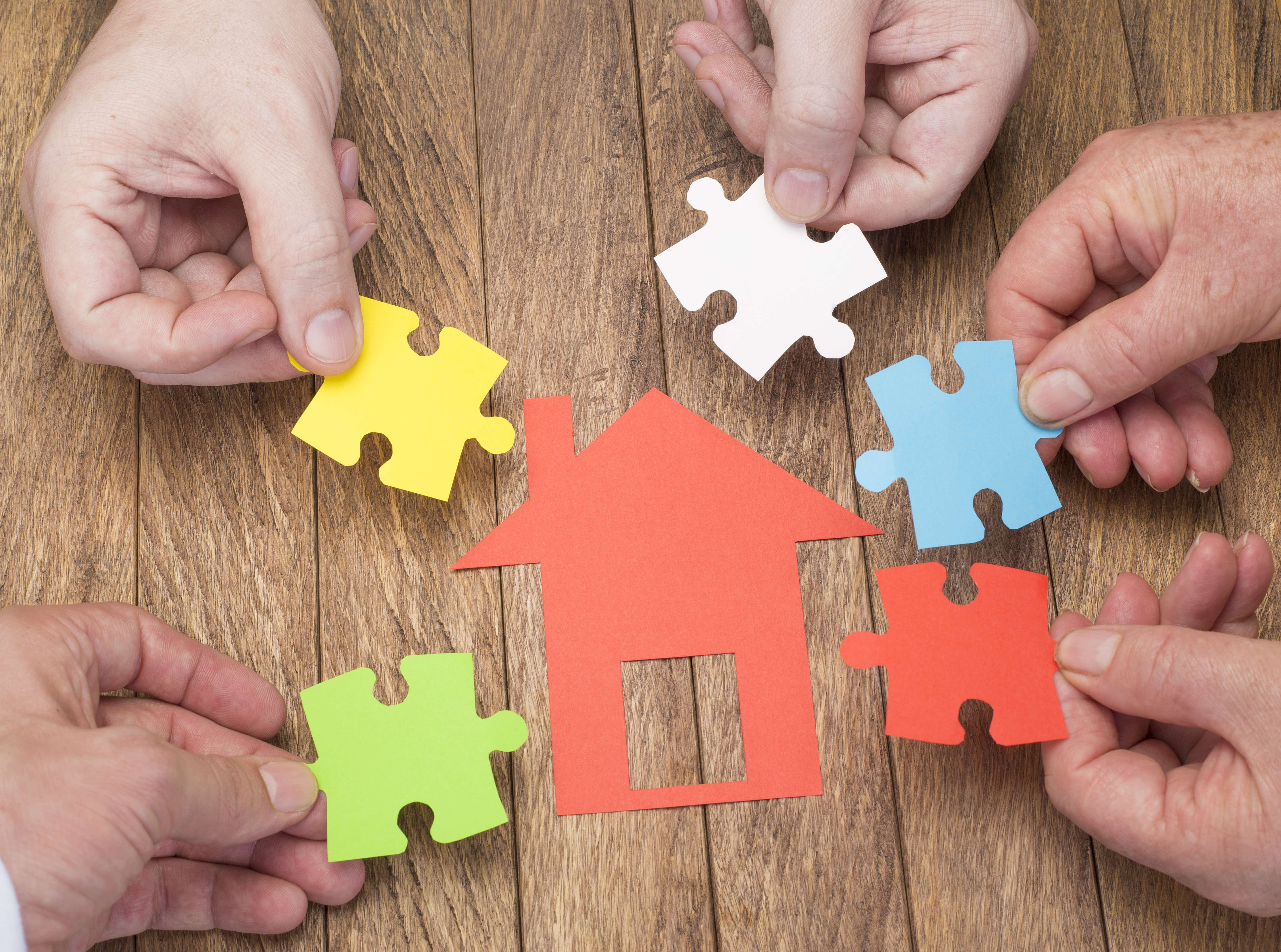 A red paper cut out of a house is laid onto a wooden desk. There are 4 hands from different people surrounding the house, each holding a paper cutout shaped like a jigsaw piece