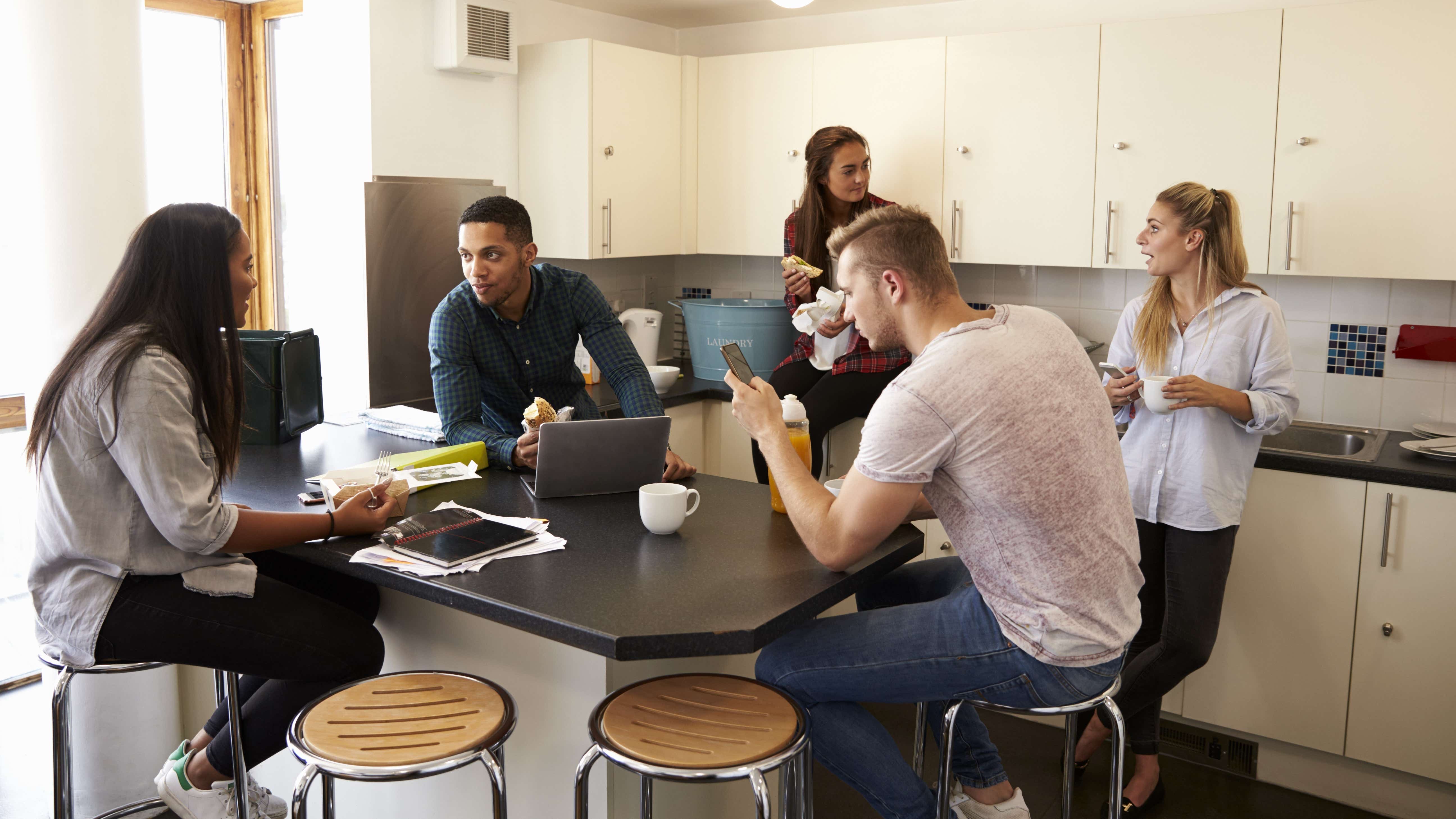 A kitchen with 5 young adults doing various things, some related to food preparation, some simply reading or chatting. This is showing a student household with a shared common area