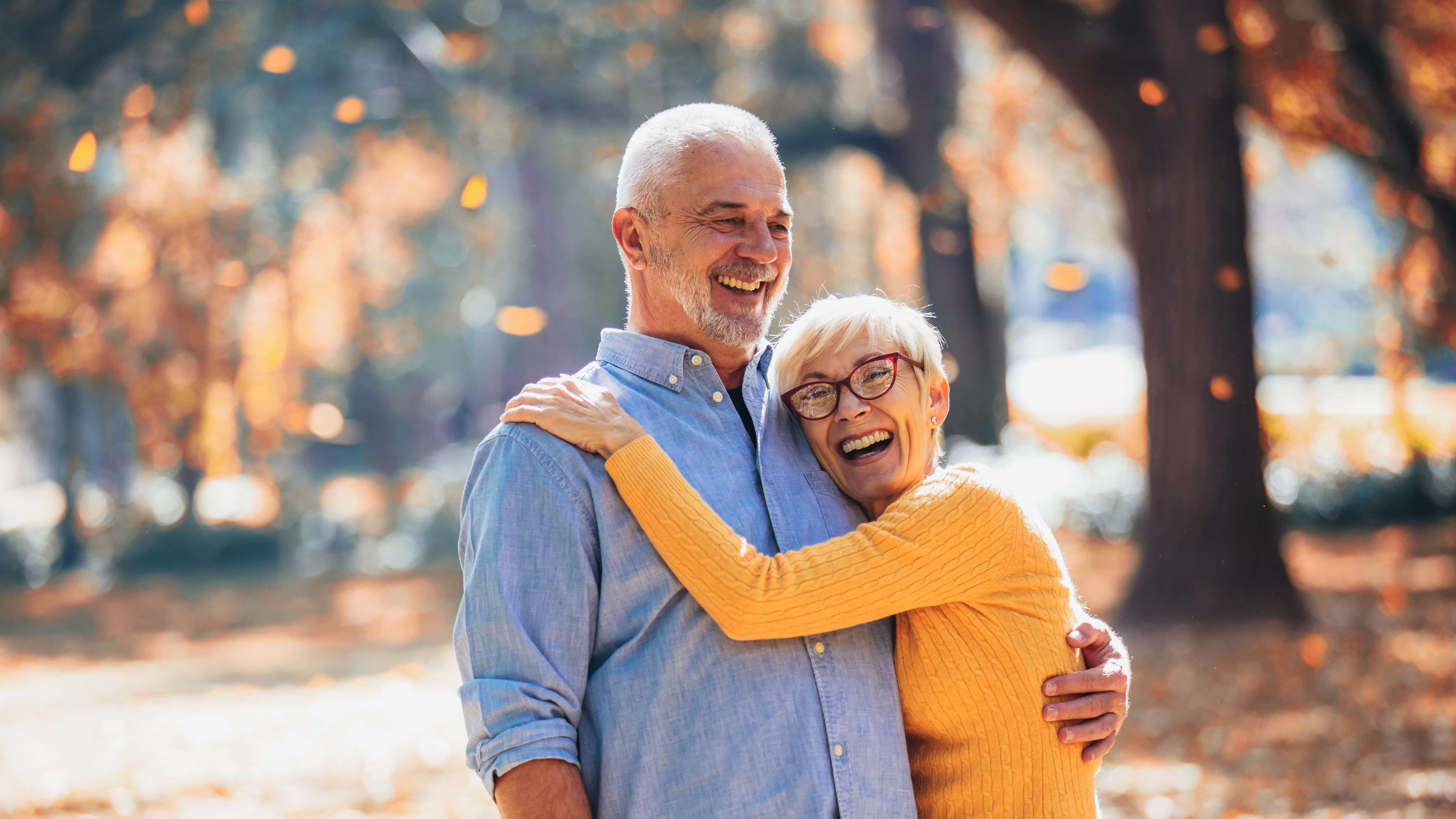 An older white male/female couple stand sideways on in front of a backdrop of autumnal trees with orange leaves falling to the ground behind them. They are laughing together