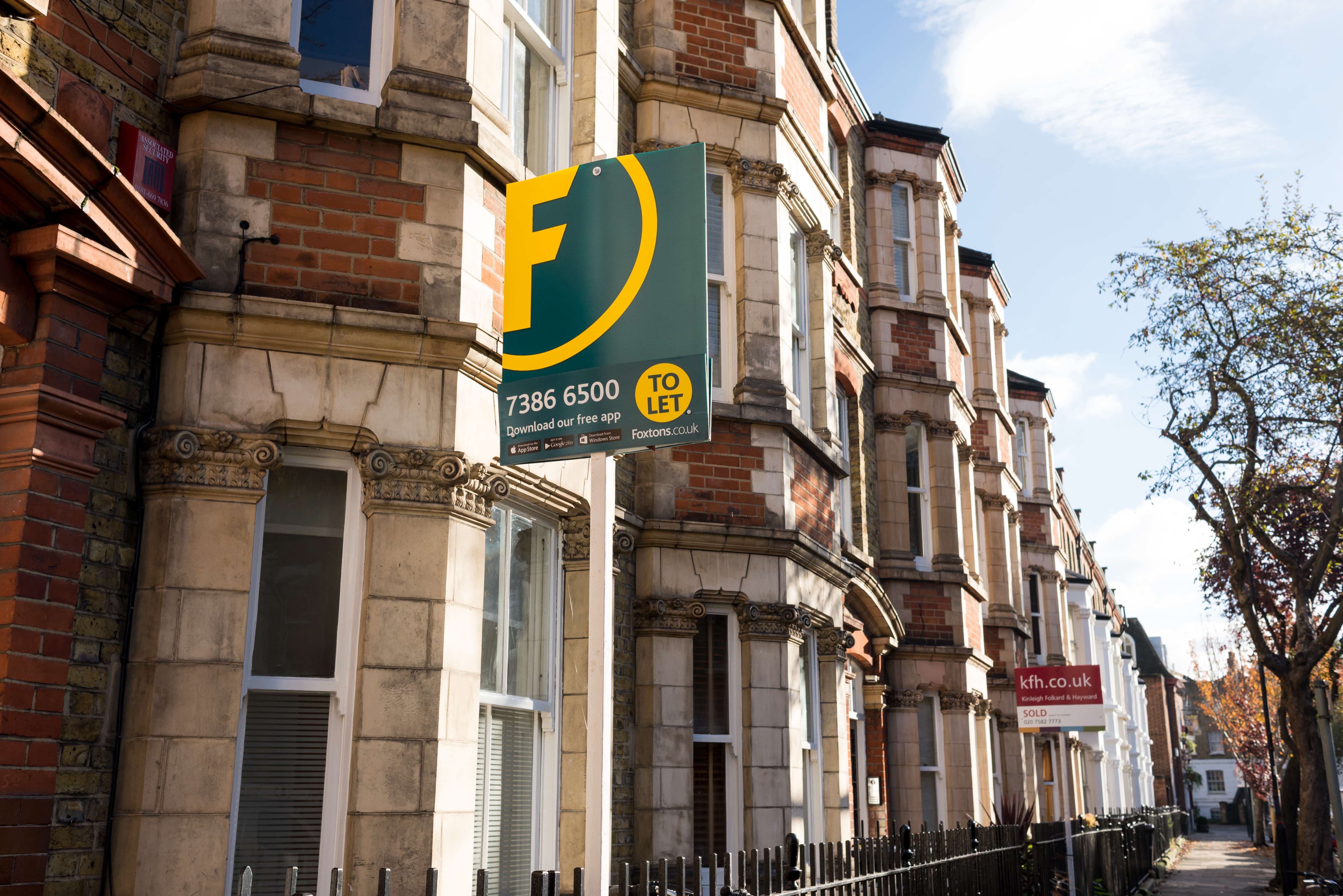 The image shows a close up of a bay window on a Victorian terraced property and there is a green to-let sign hanging from just above the window. There are also a row of further terraced houses, some pavement and some trees in the background