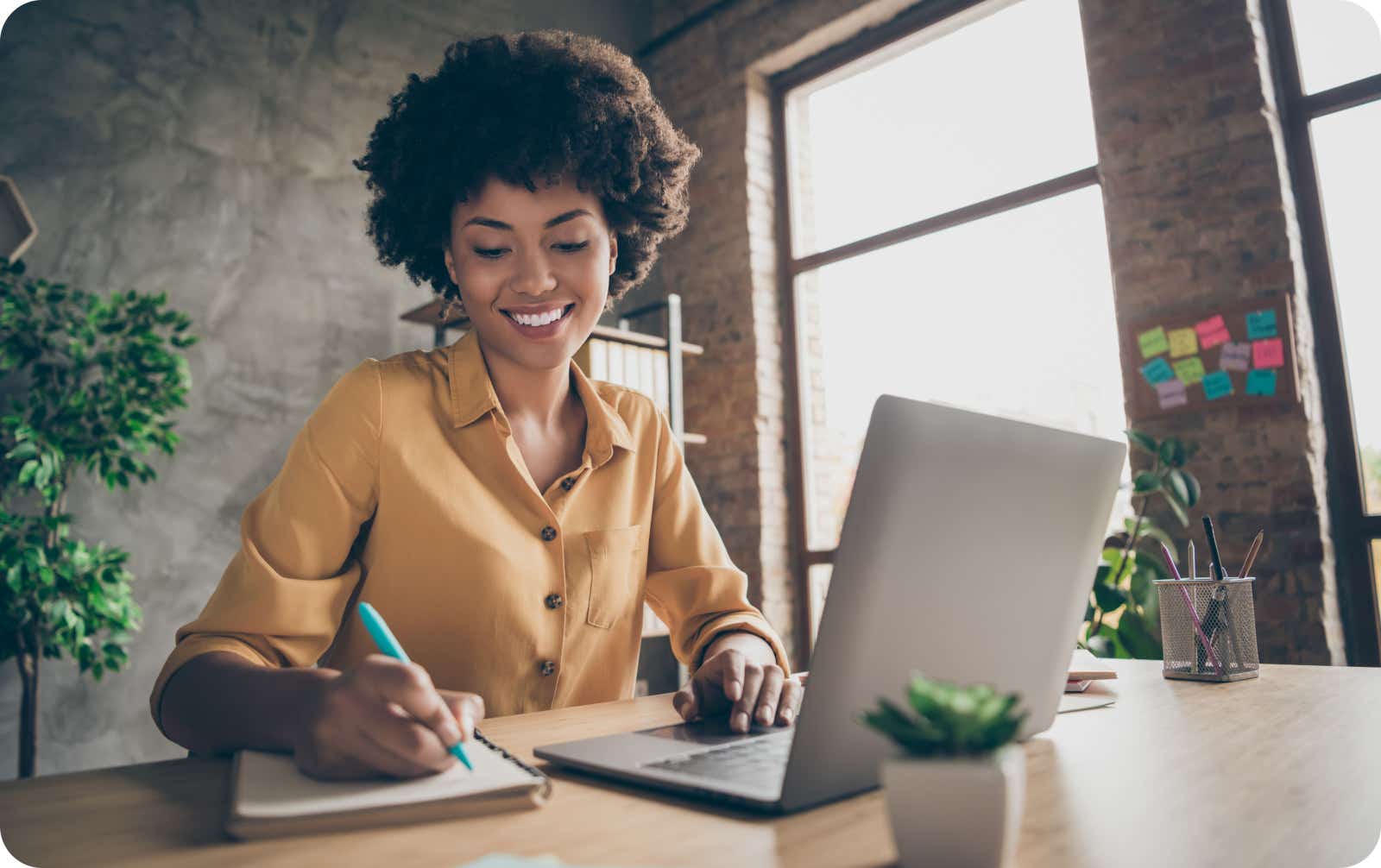 Woman working at a desk writing on a notepad and looking at a laptop