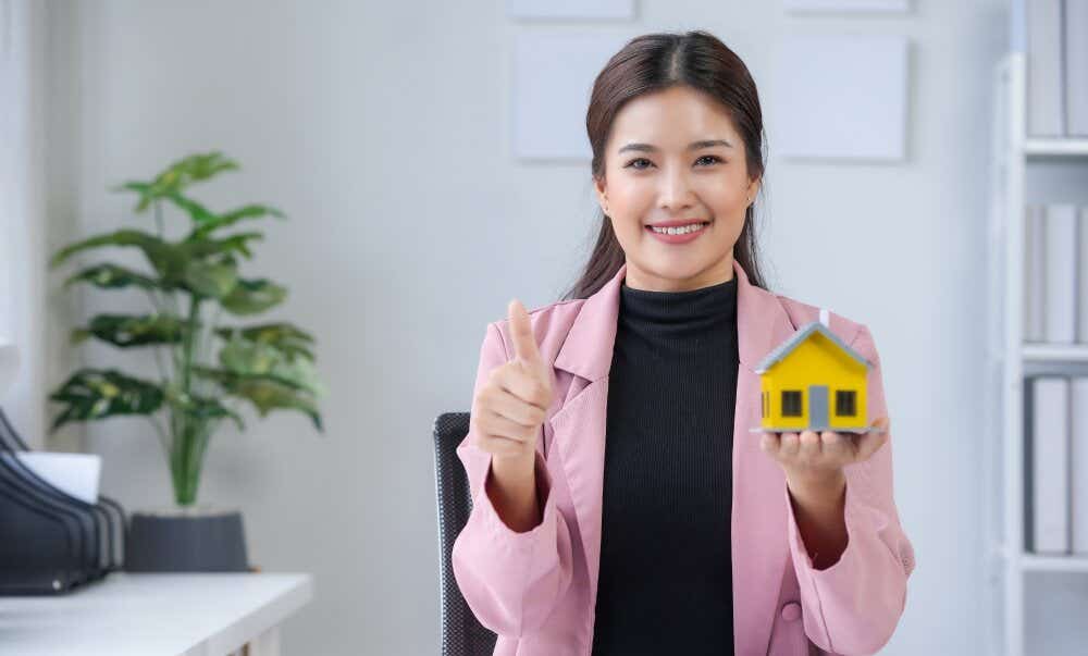 An image of a smiling woman holding a model home in her right hand while doing the 'thumbs up' sign with her left.