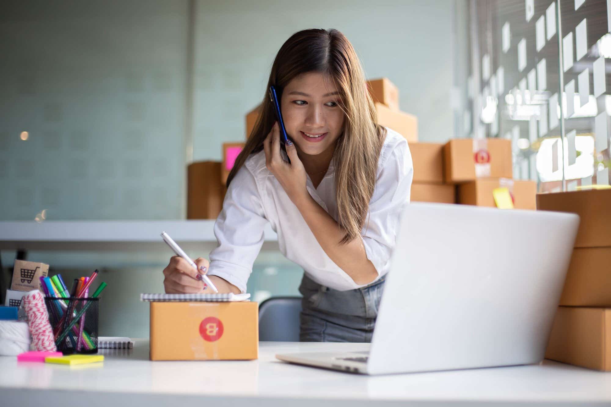 Small business owner on the phone, writing down a message and looking at her laptop.