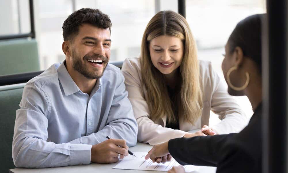 An image of a smiling couple discussing a house purchase with an advisor in an office.