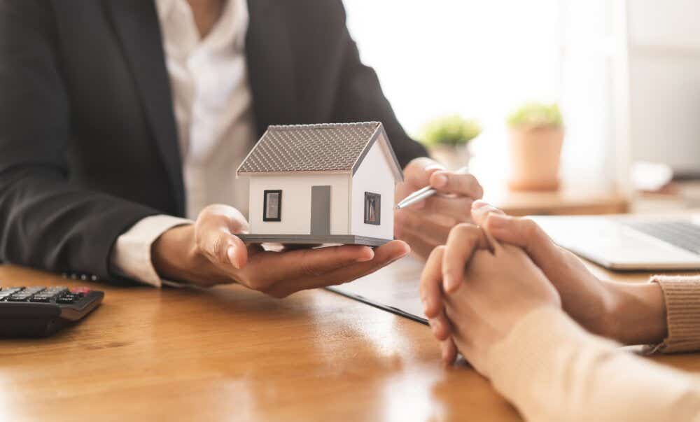 An image of a professional man holding a model home in an office while talking to a client.