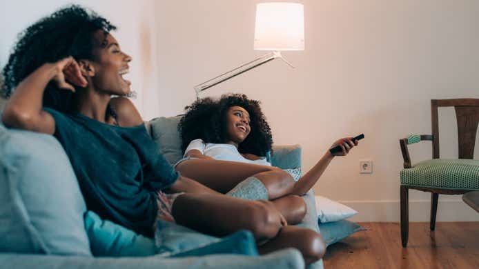 Happy young two black women lying down on the couch watching tv