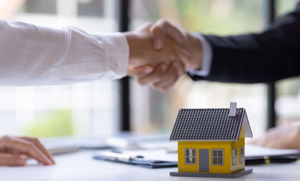 An image of two people shaking hands in an office alongside a model home and a contract