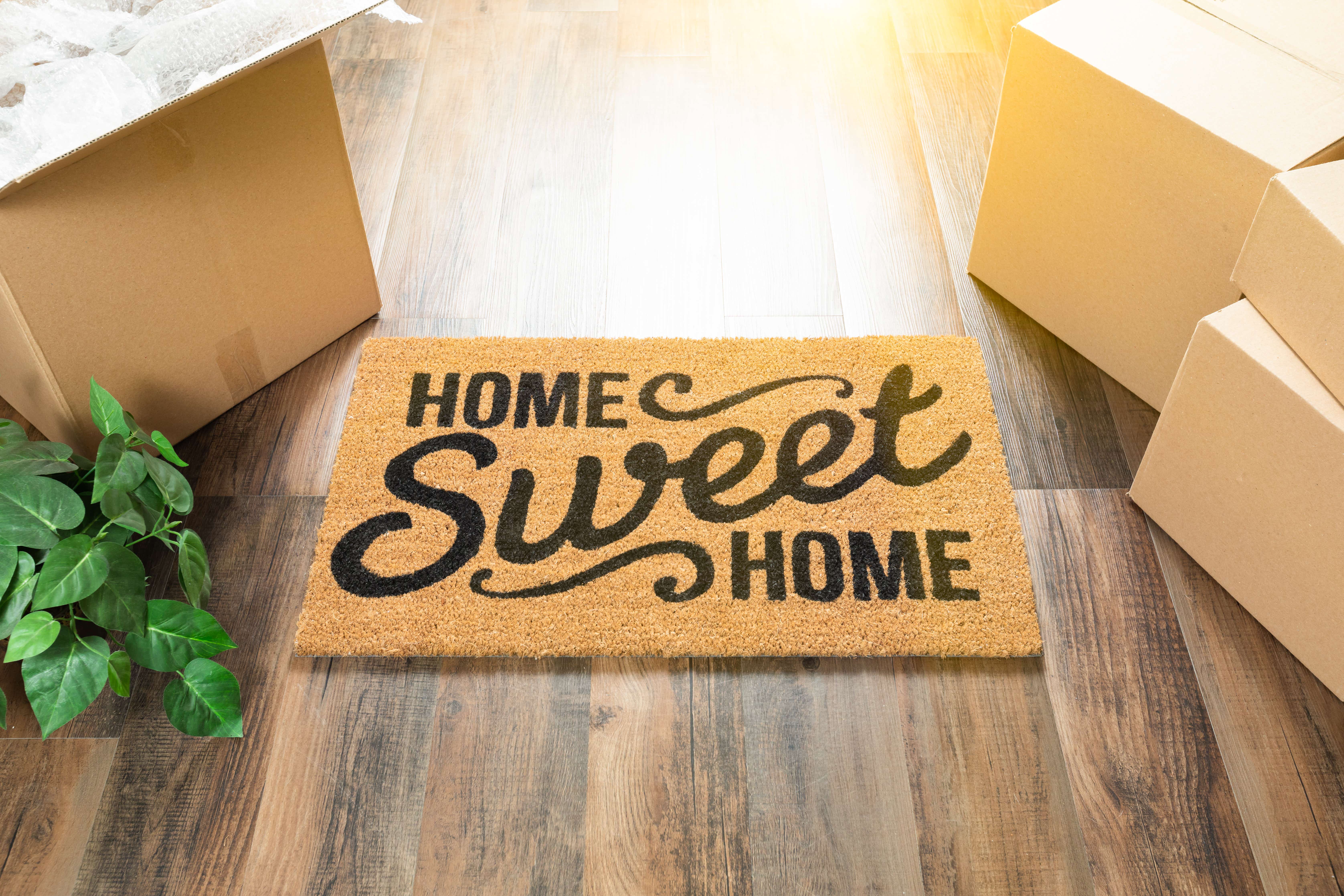 A hessian door mat which has the words 'Home sweet home' printed on it sits on the floor of a sunny hallway, surrounded by cardboard boxes and a house plant