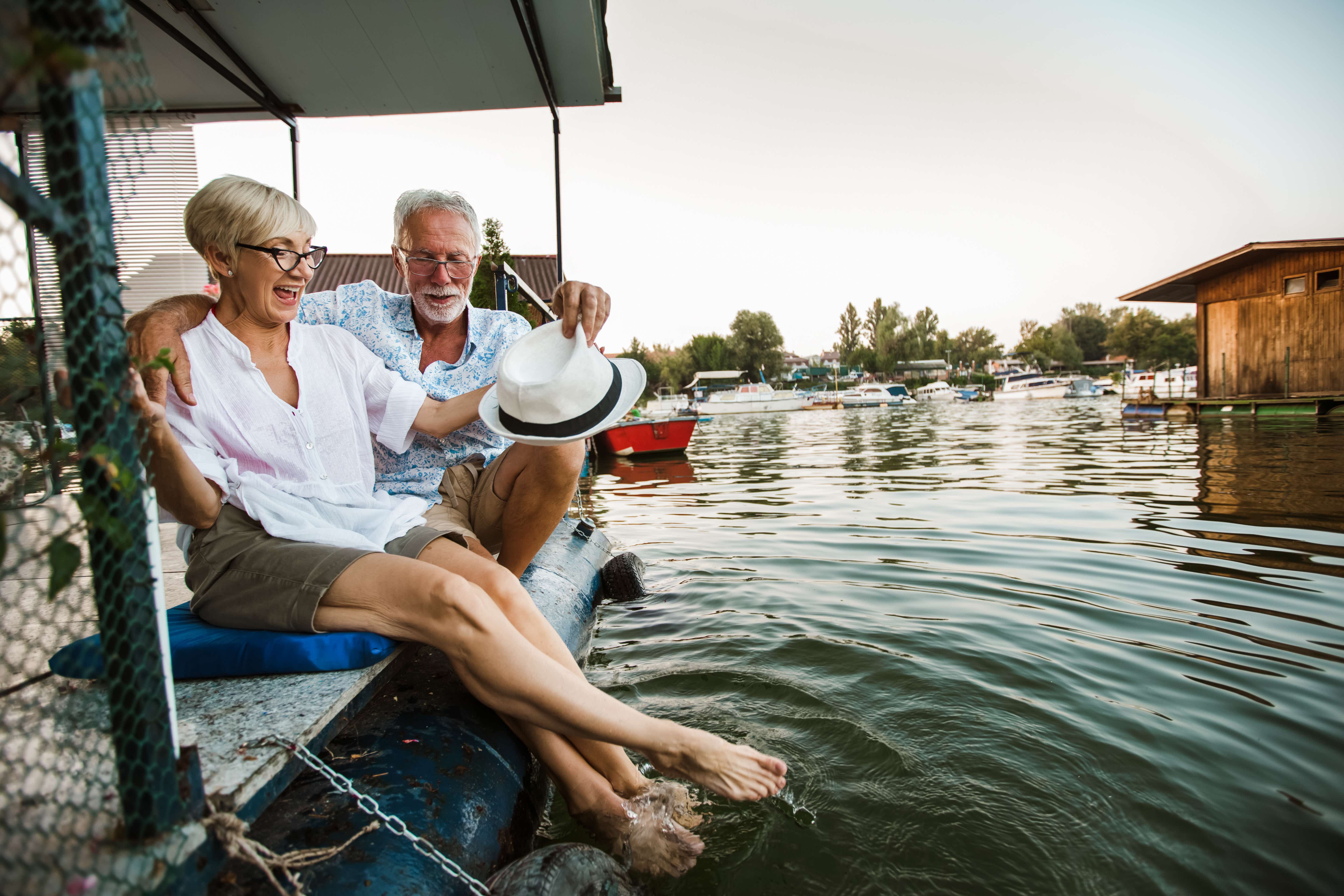 An older male/female couple sits on the side of a boat dipping their feet in the water and smiling. The boating lake continues into the background with a wooden hut and trees along the horizon.
